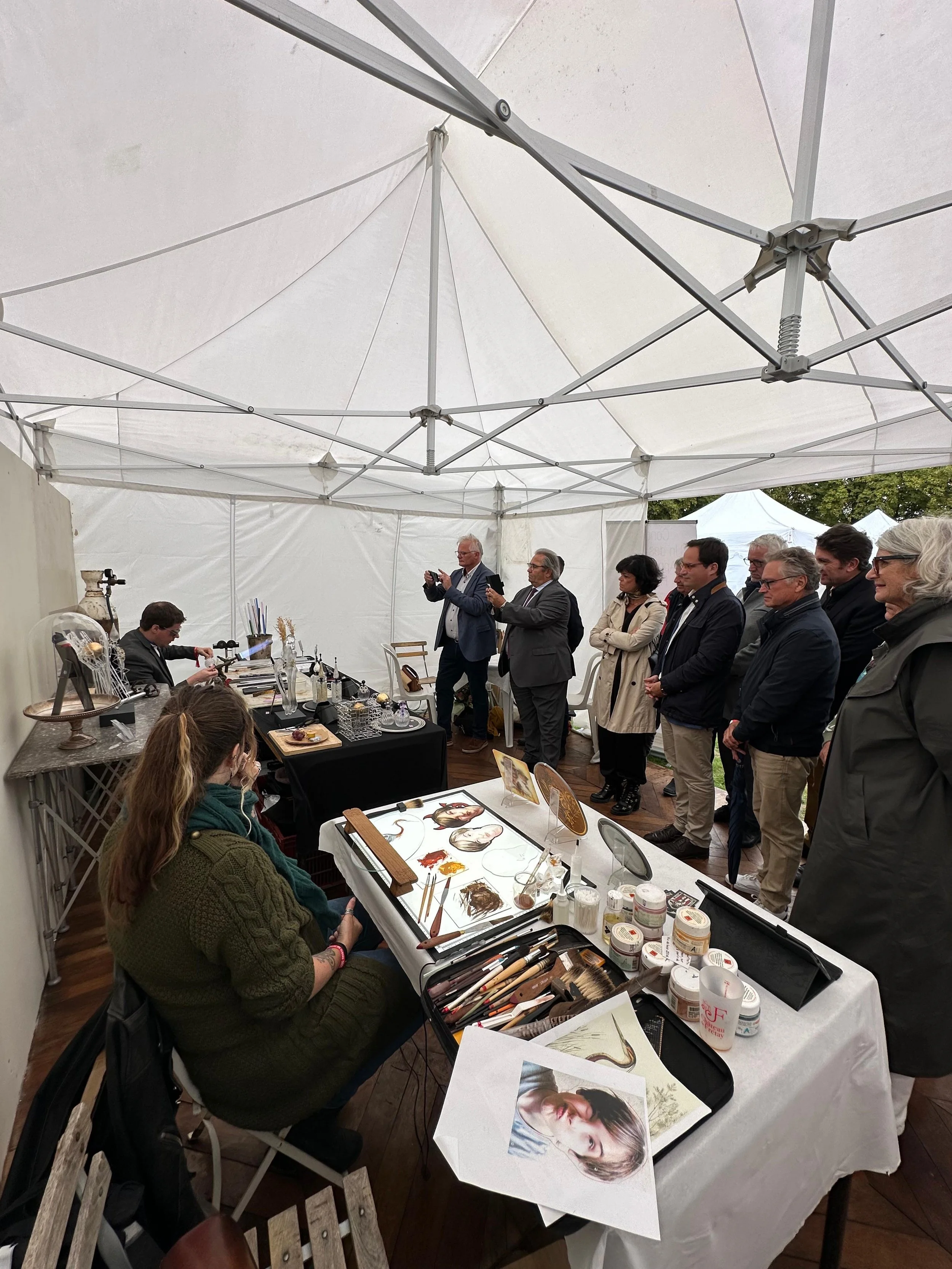 Une artiste réalise une peinture lors d'une exposition sous une tente blanche, avec plusieurs personnes observant et prenant des photos.