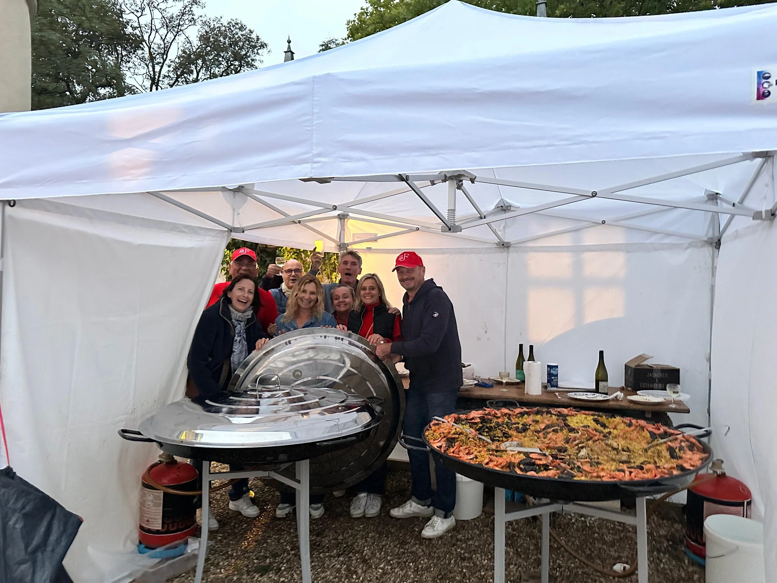 Groupe de personnes souriantes à une fête, devant une grande paella et un plat de fruits de mer, sous une tente blanche.