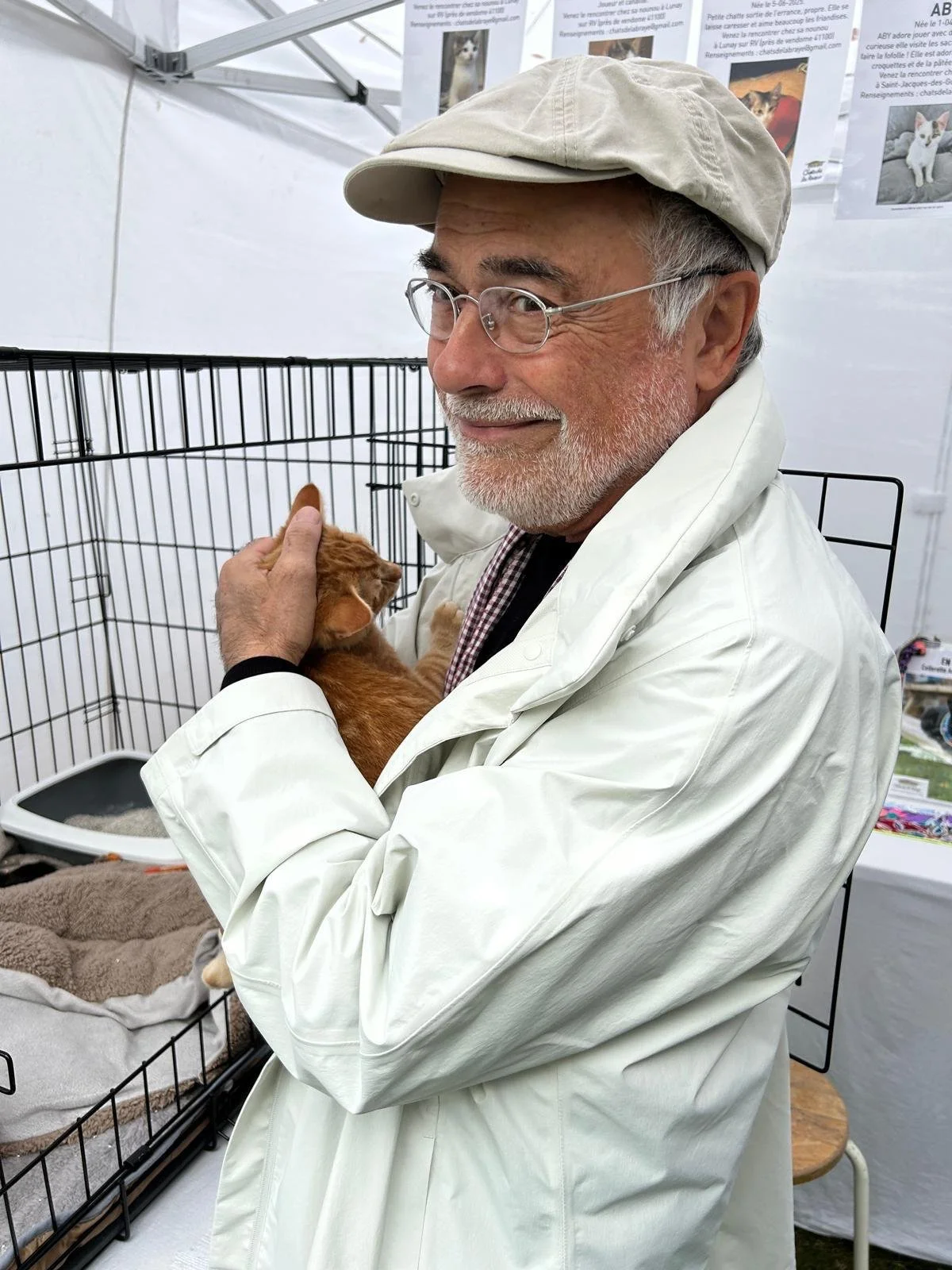Un homme âgé portant des lunettes, une casquette beige et un manteau blanc, tient un chat orange dans ses bras à l'intérieur d'une cage en métal.