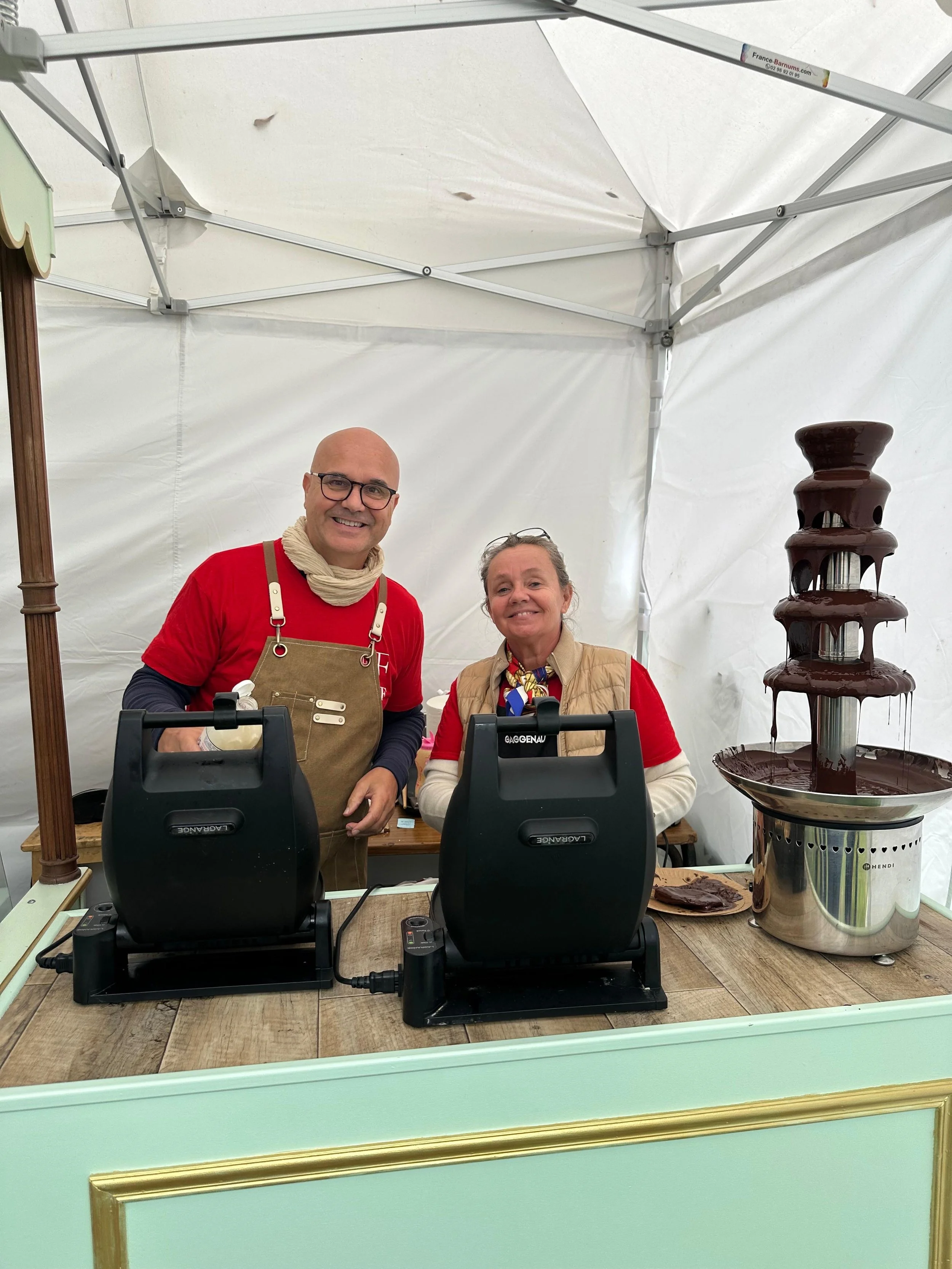 Deux personnes souriantes derrière un stand de crème glacée ou chocolat chaud, avec une fontaine de chocolat en dessert à côté.