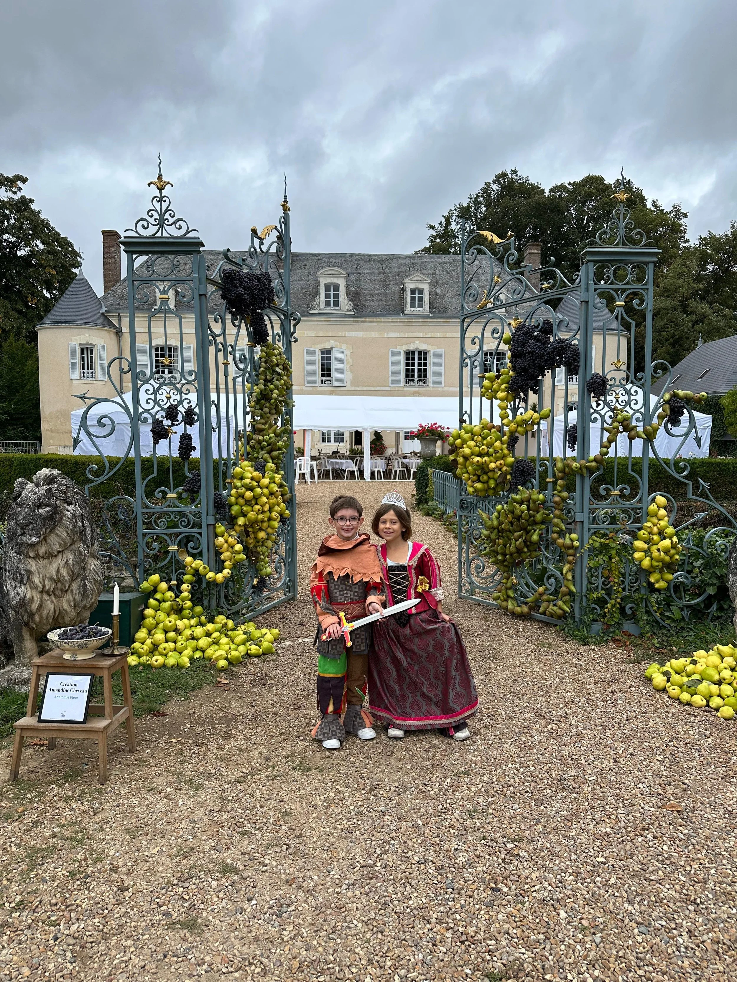 Deux enfants déguisés en chevaliers et en princesses posent devant une entrée de château ornée de pommiers, avec un bâtiment en arrière-plan lors d'une fête médiévale.