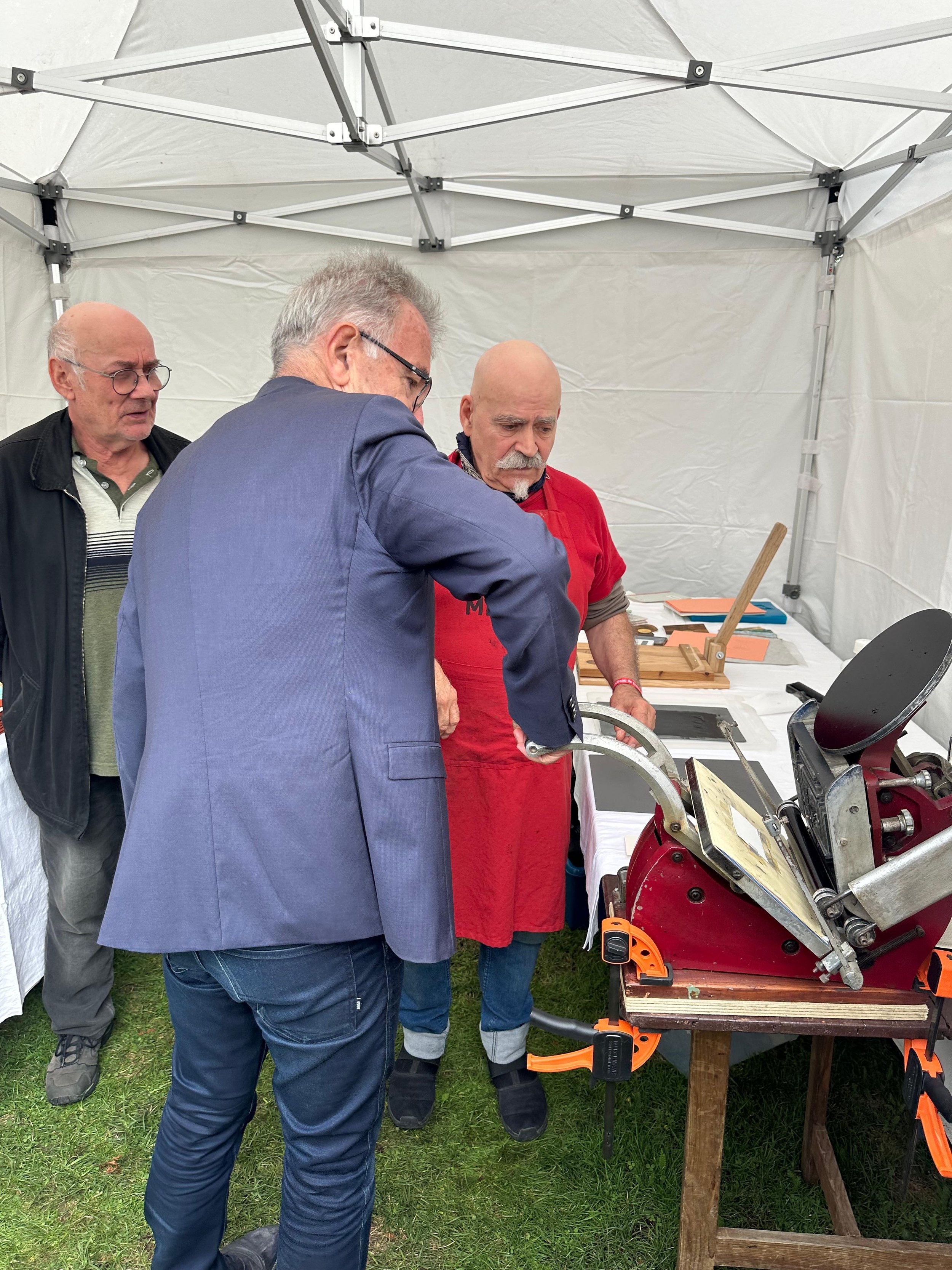 Trois hommes dans une tente discutent autour d'une machine ancienne à usage artisanal, posée sur une table en bois.