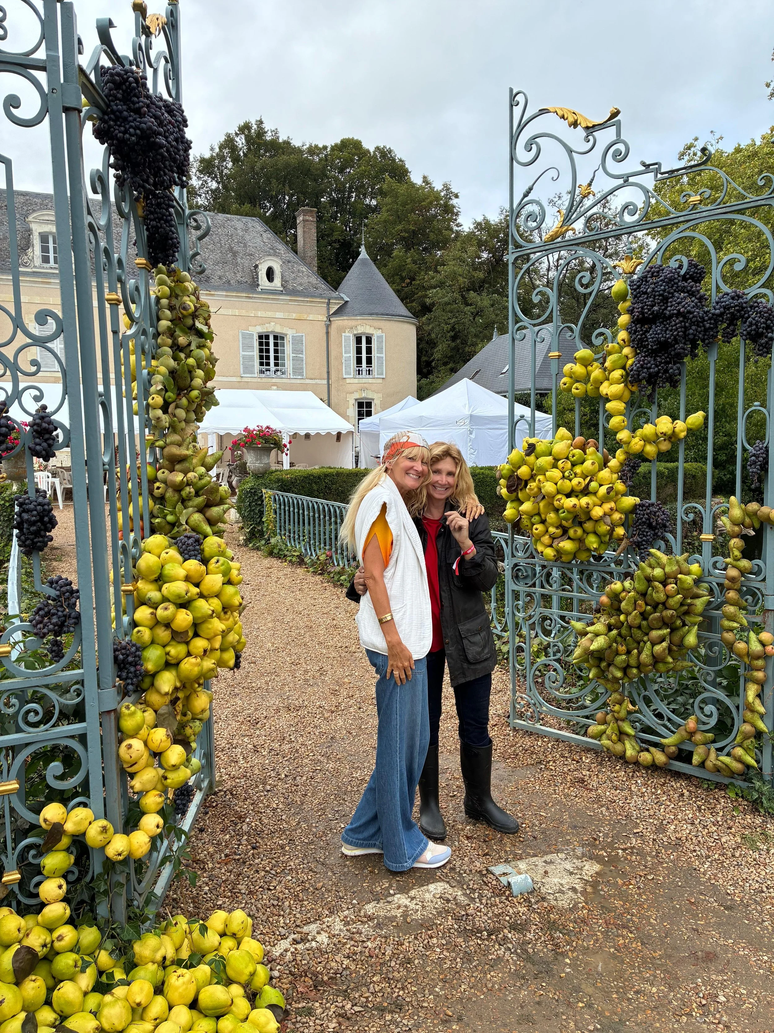 Deux femmes souriantes posant dans une entrée ornée de grappes de raisins et de pommes, devant un château en pierre avec des tentes blanches en arrière-plan.
