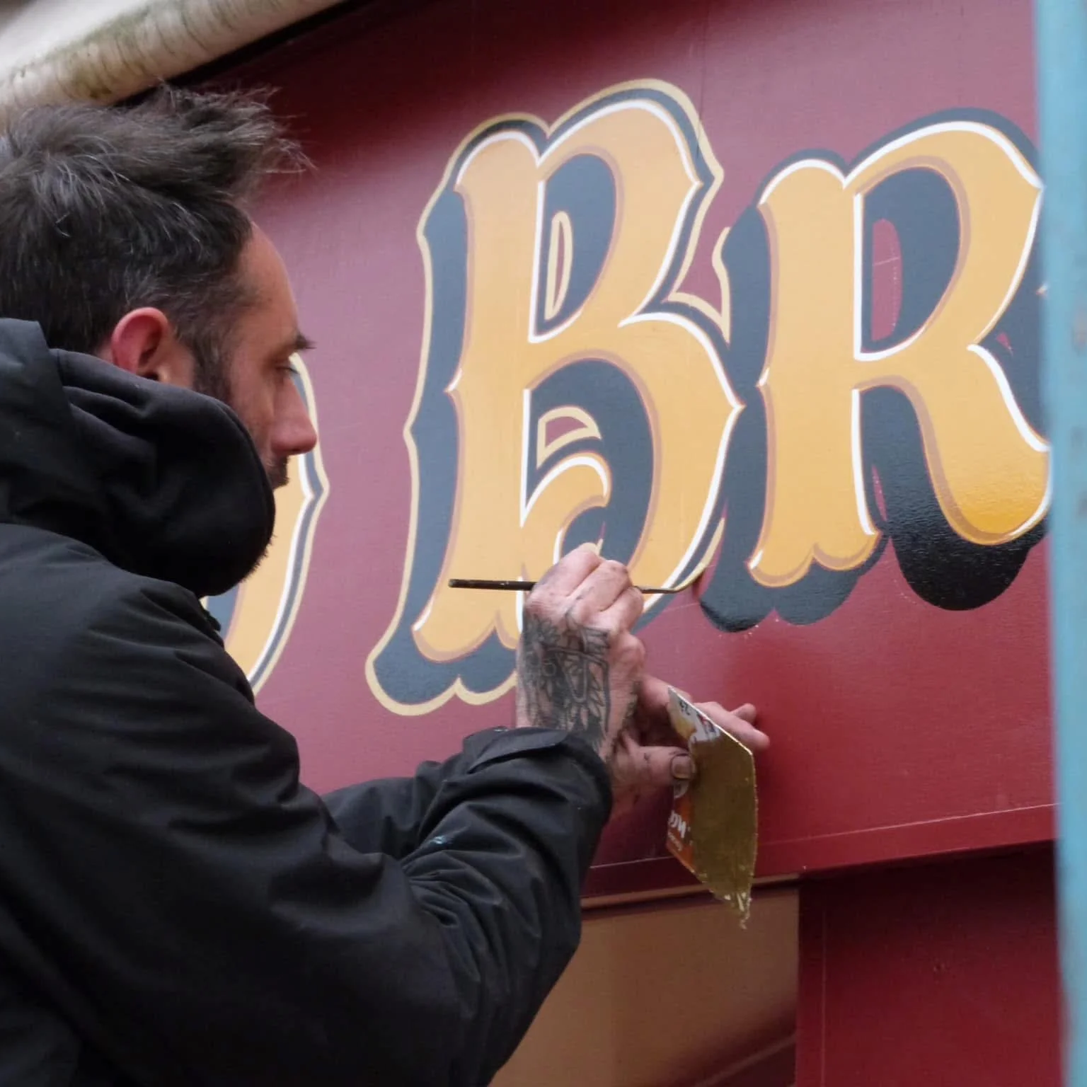 Un artiste est en train de peindre un panneau en utilisant des couleurs dorées et noires pour créer des lettres stylisées sur un fond rouge.