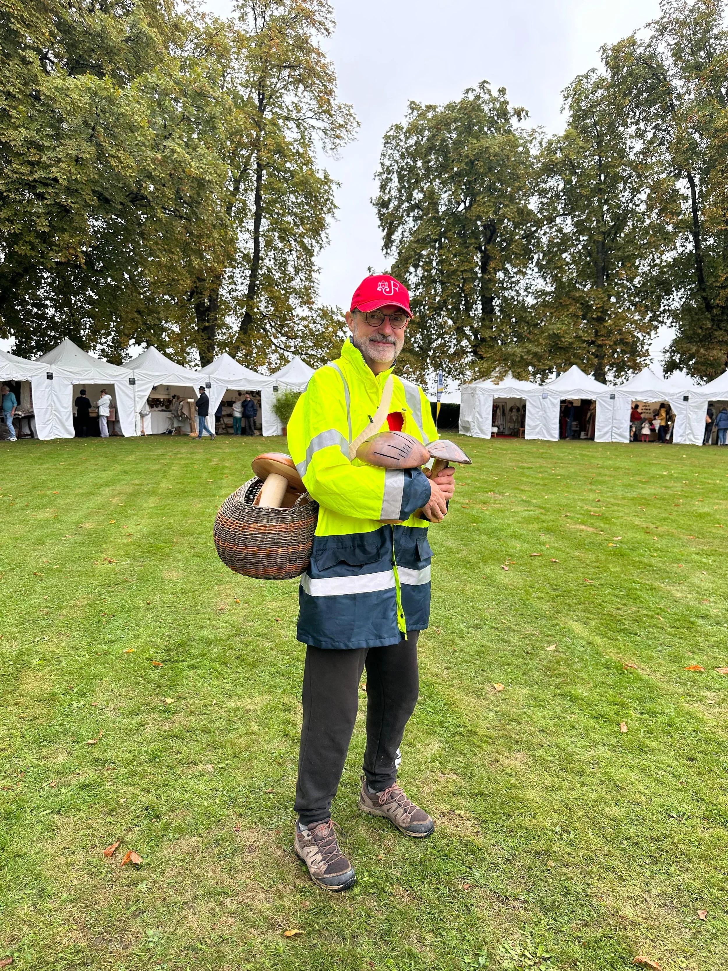 Un homme portant un manteau jaune fluorescent et un chapeau rouge se tient dans un parc avec des stands de tentes blanches en arrière-plan, tenant une sculpture en bois.