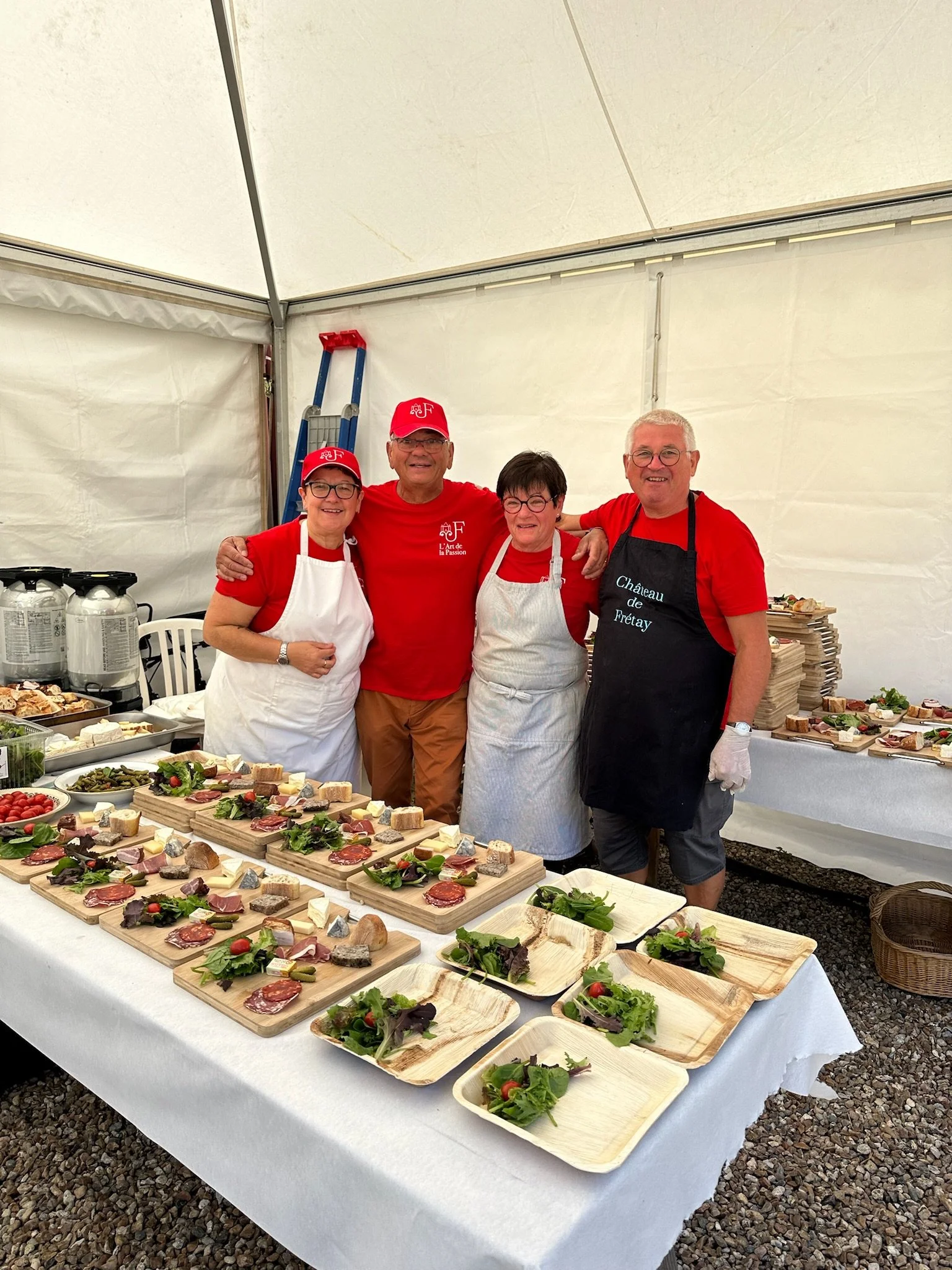 Groupe de quatre personnes souriantes en vêtements rouges et blancs, posant derrière un buffet de plats froids avec des toasts, salades et garnitures, dans une tente blanche.