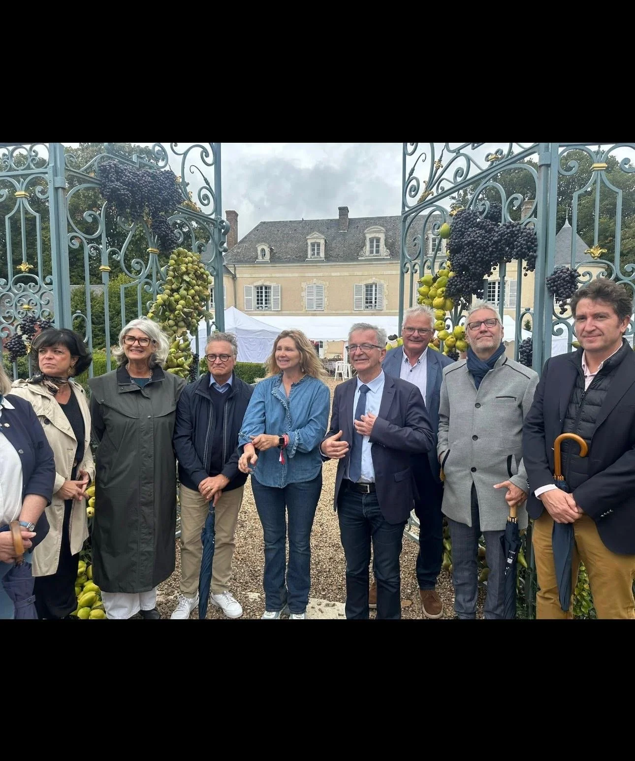Groupe de personnes posant devant une porte ornée de raisins et de grappes de raisin, avec une bâtisse ancienne en arrière-plan, lors d'un événement en plein air par temps nuageux
