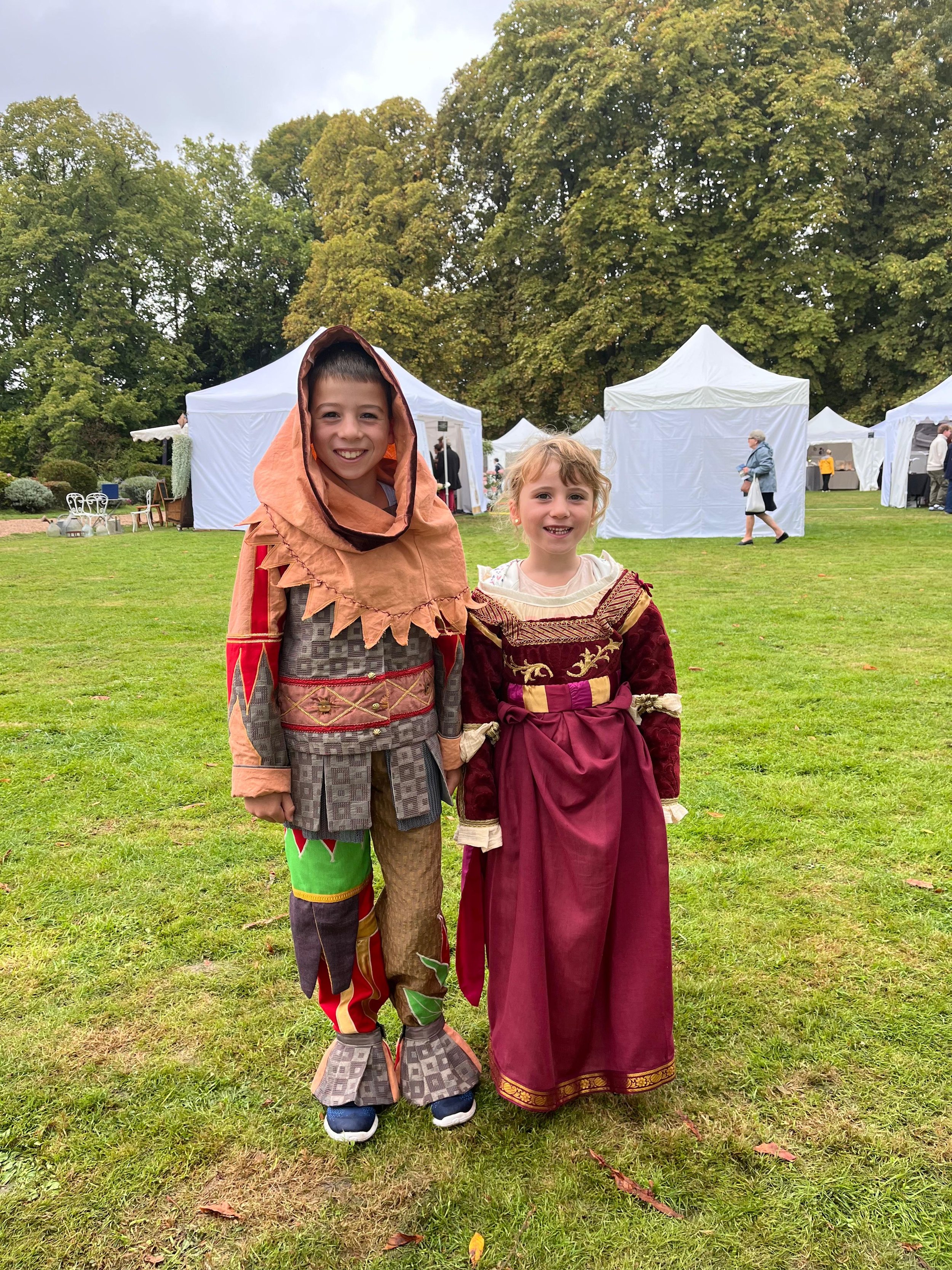 Deux enfants en costumes médiévaux, un garçon avec une tunique colorée et un capuchon, une fille en robe rouge et dorée, debout sur un terrain de festival avec des tentes blanches en arrière-plan, des arbres et des personnes en promenade.