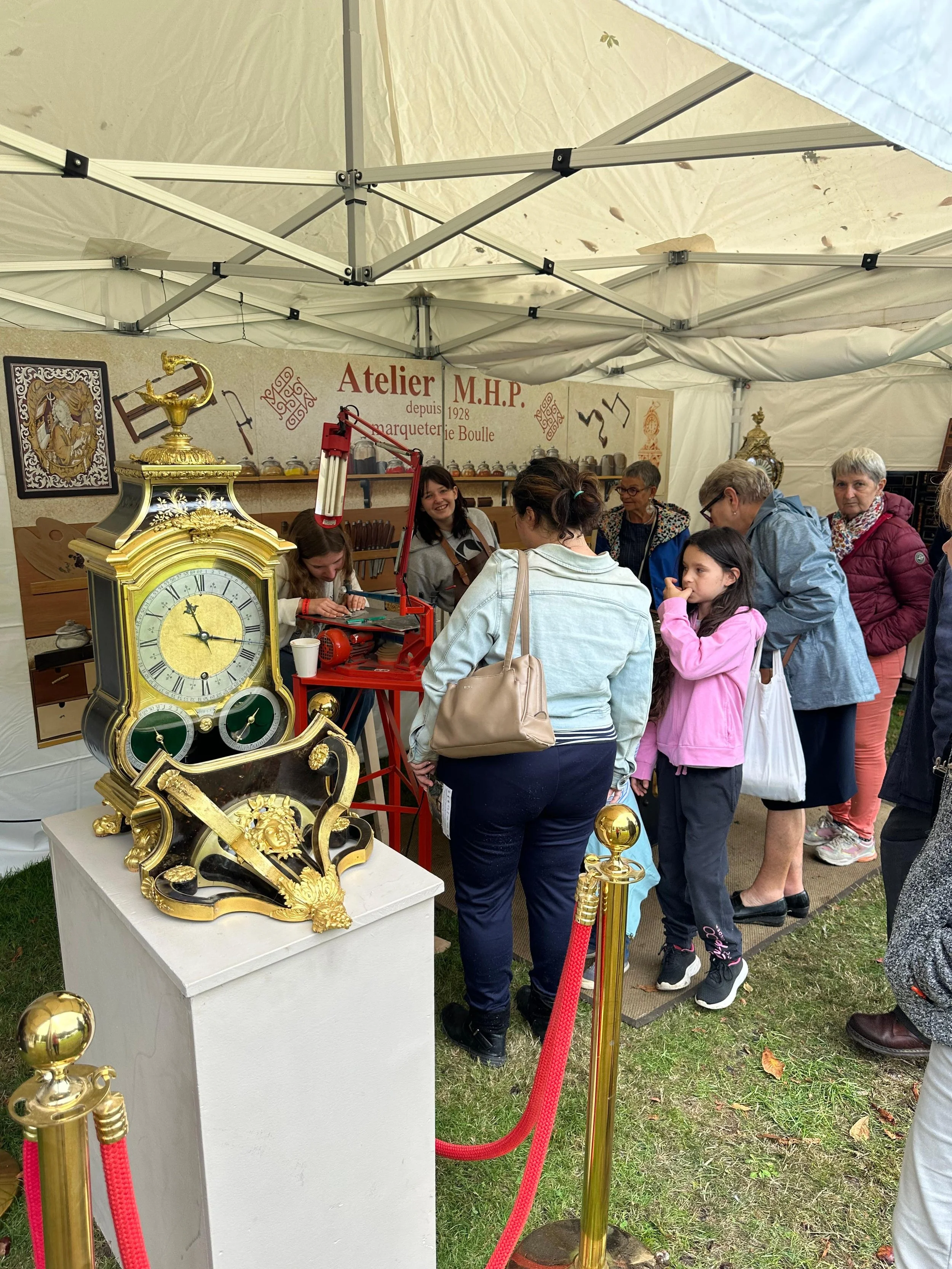 Des gens regardent une exposition d'horlogerie dans une tente, avec une horloge ornée de dorures sur un piédestal devant eux.