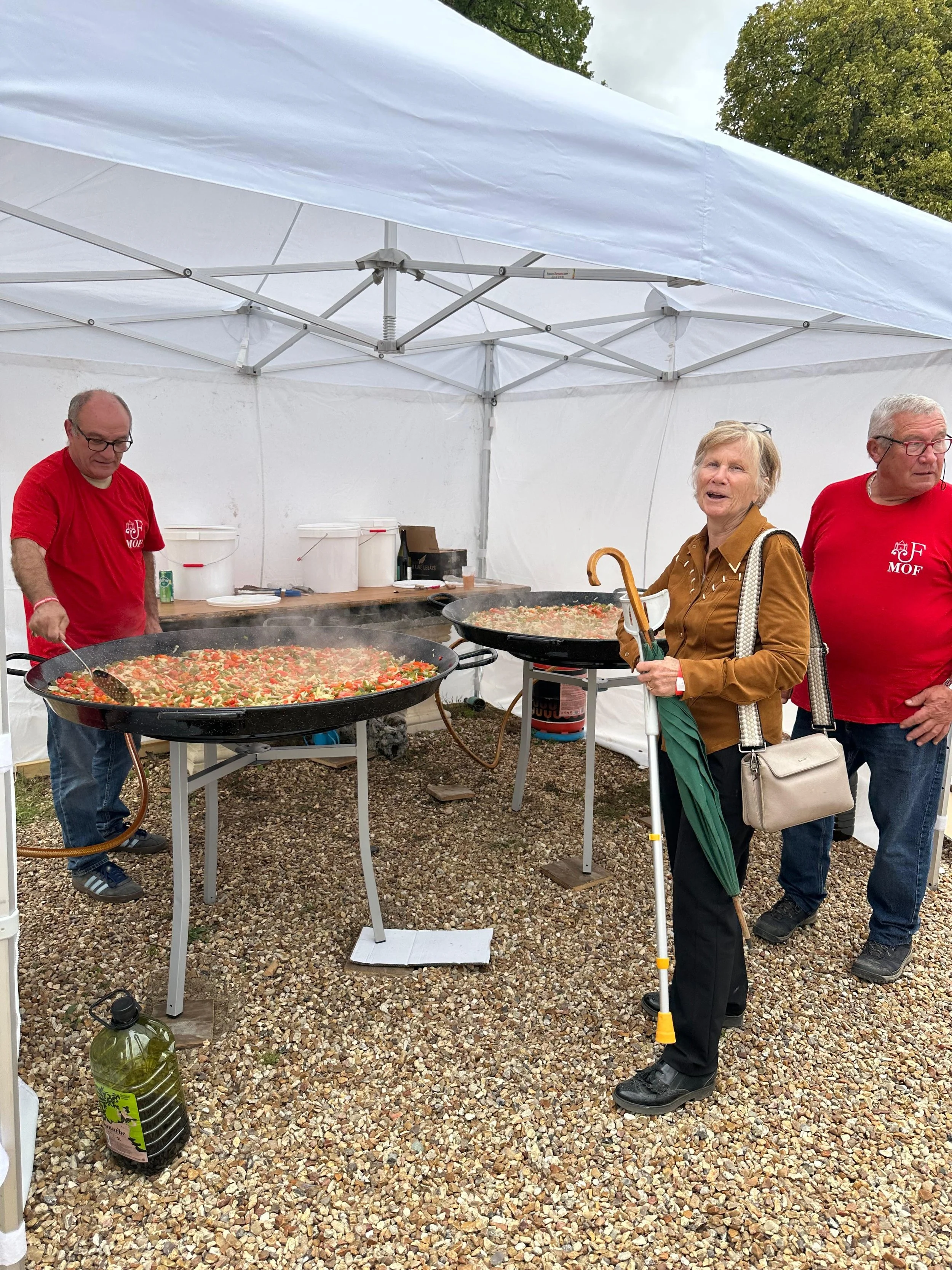 Trois personnes, deux hommes et une femme, dans une tente en plein air, cuisinant des paellas dans deux grands râteaux à paella. La femme porte une canne. Au fond, une table avec des conteneurs et des ustensiles.