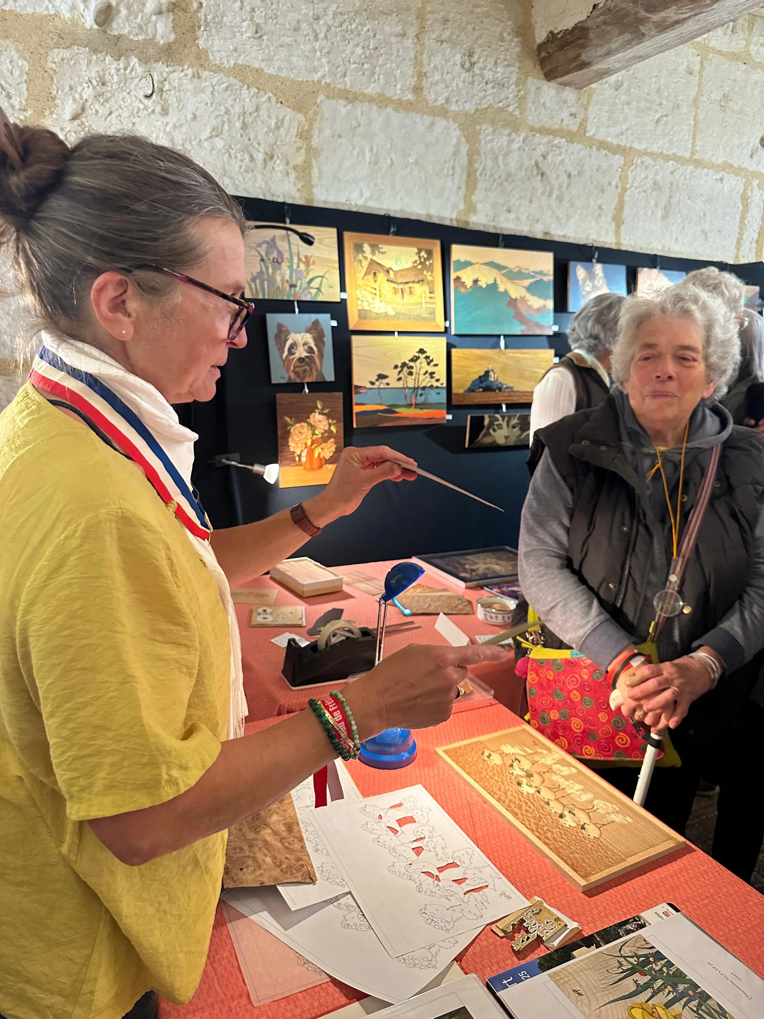 Une femme en jaune utilisant un pointeur lors d'une démonstration ou d'une présentation d'artisanat, avec deux autres femmes à côté. Des peintures sur toile sont exposées en arrière-plan.
