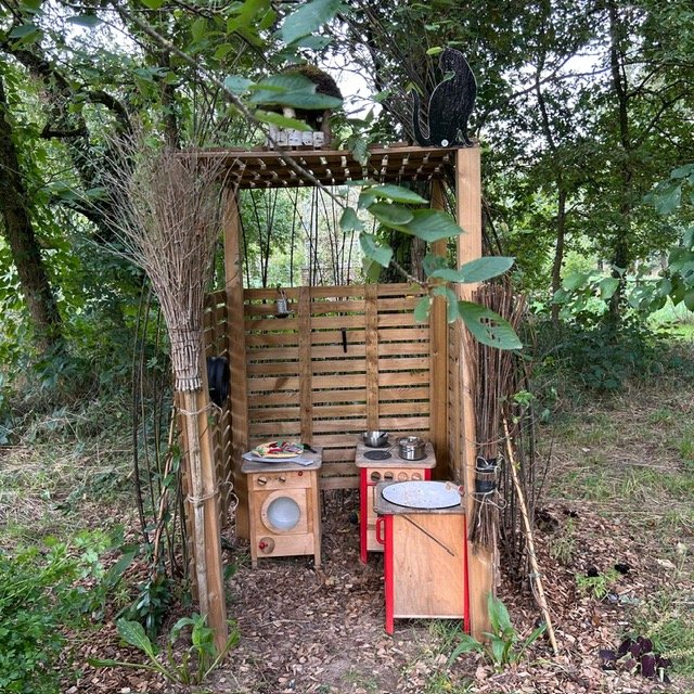 Petite cabane en bois en plein air, décorée avec des objets en terre cuite, plantes vertes et décorations, entourée de végétation.