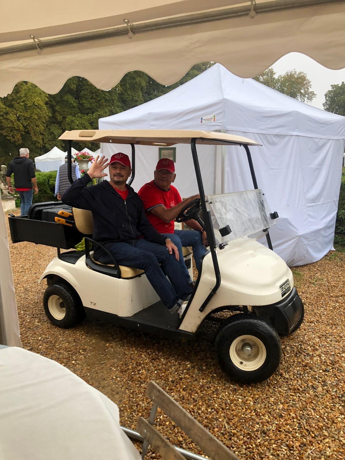 Deux hommes assis dans une voiturette de golf, l'un salut à la caméra, dans un environnement extérieur avec des tentes blanches, un chemin en gravier et des arbres en arrière-plan.