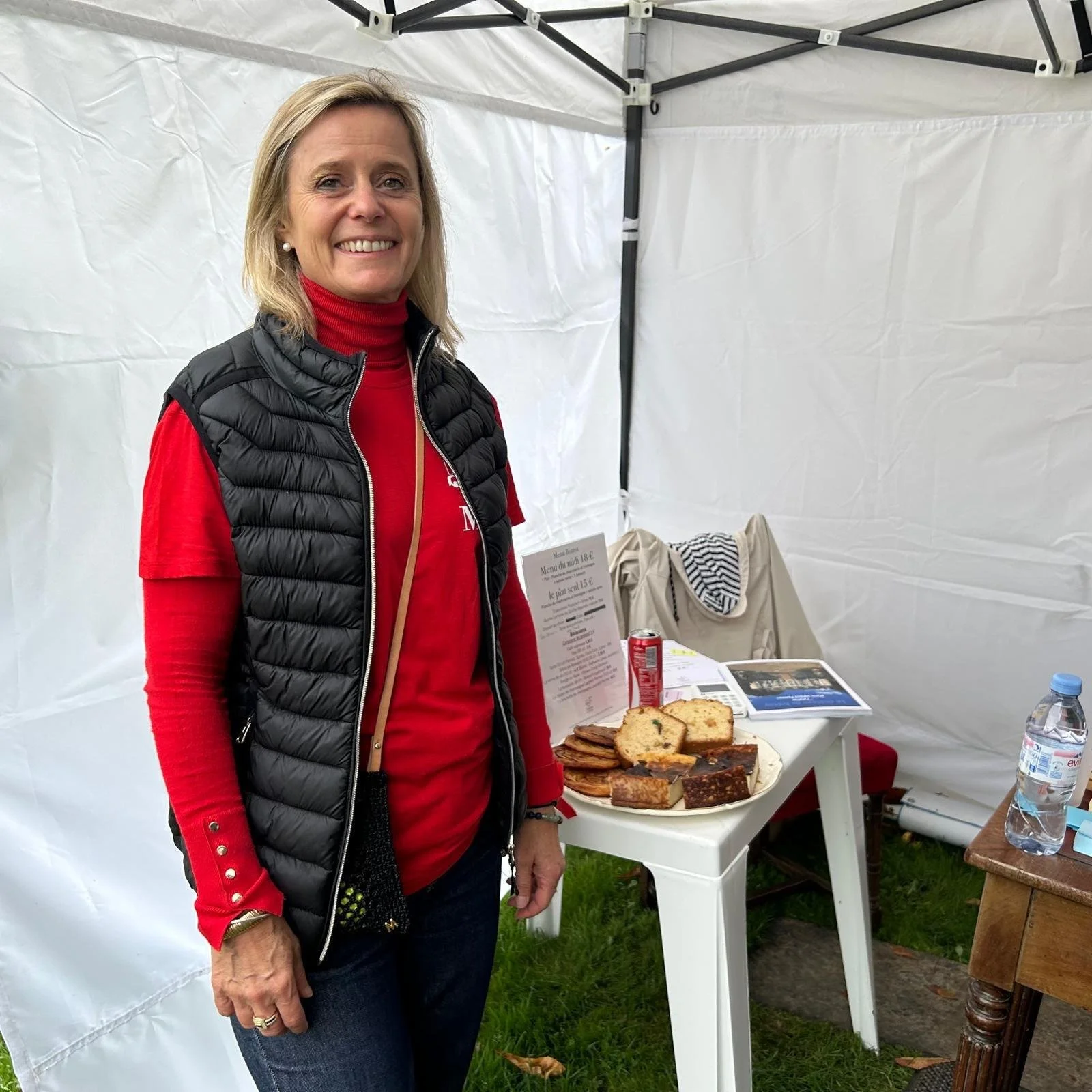 Une femme souriante debout à côté d'une table avec des cakes, une canette, une bouteille d'eau et des documents dans un stand en tissu blanc.