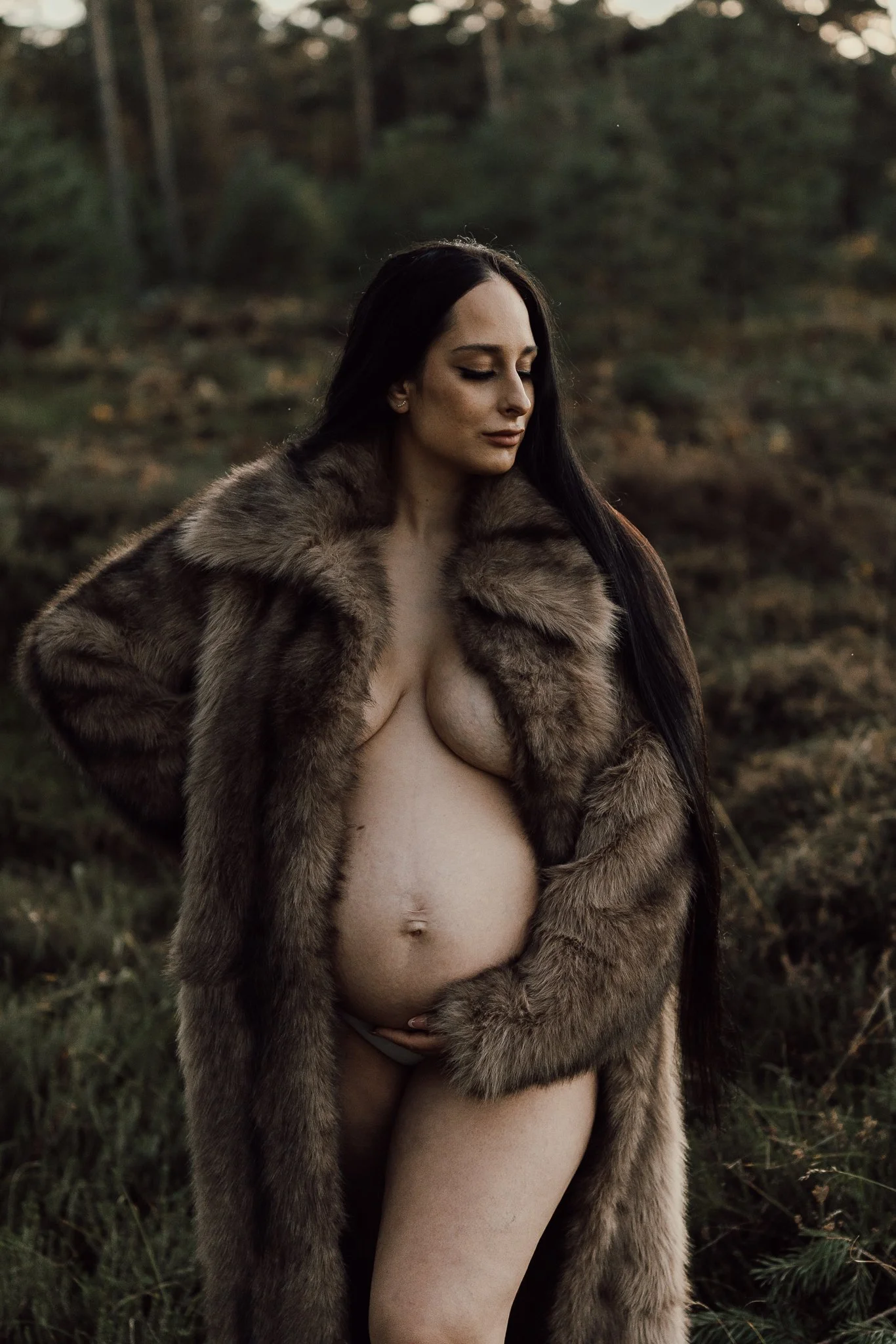 Gravid kvinna med långt mörkt hår står i ett högt gräslandskap, svept i en stor pälsrock som framhäver hennes gravidmage.