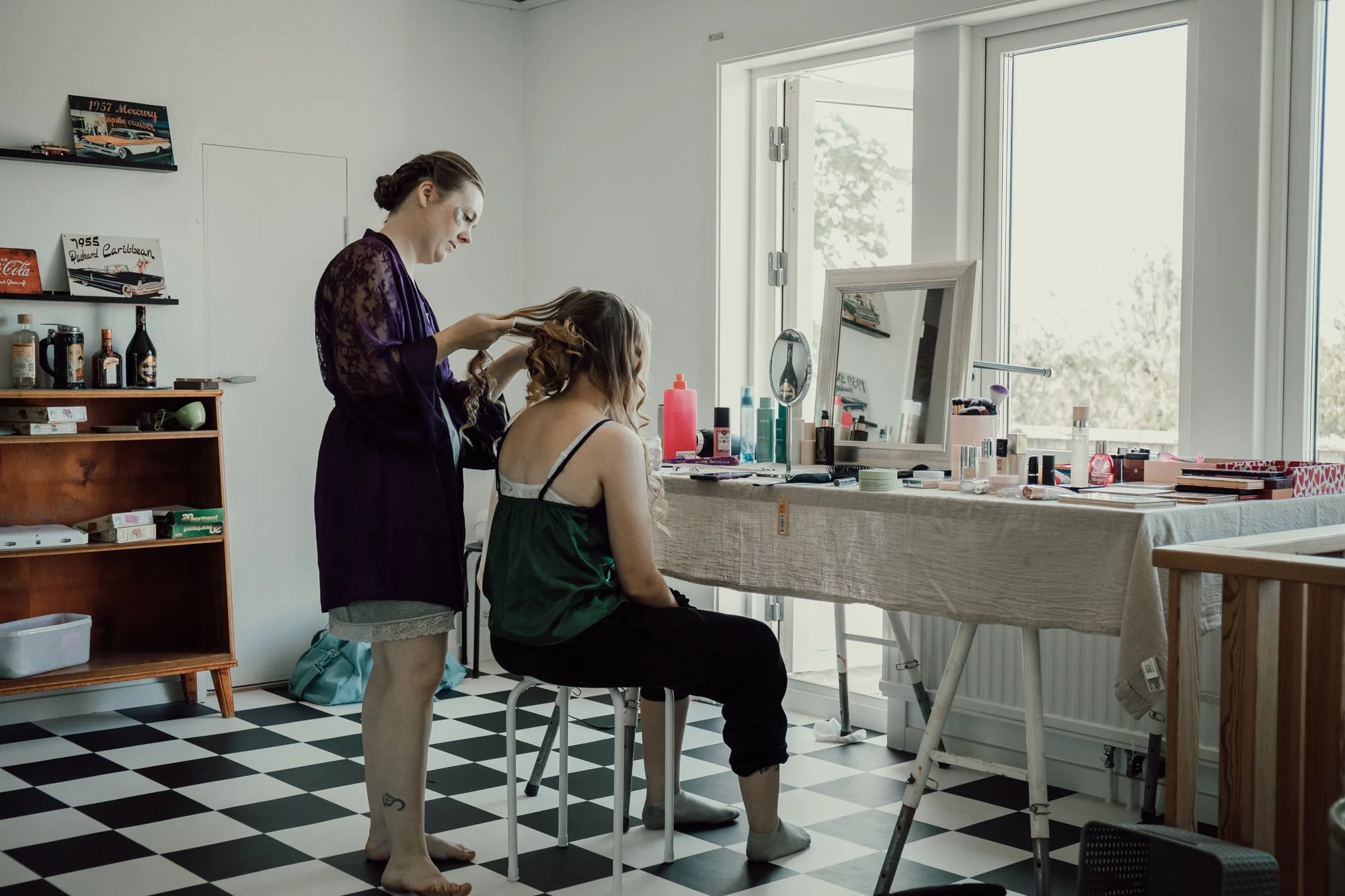 Tjej sitter på en pall framför ett sminkbord och en annan tjej står bakom och lockar hennes hår.