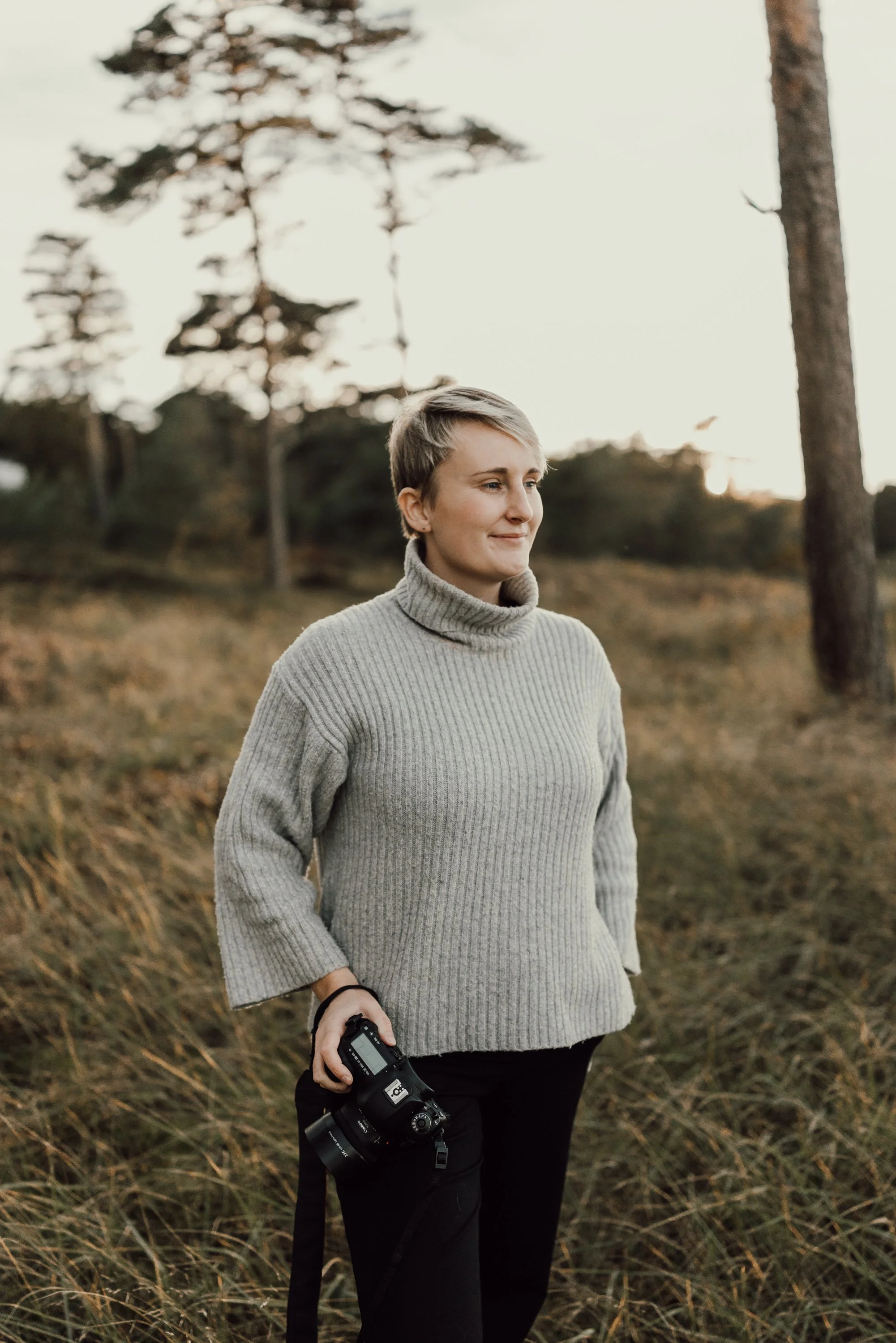 Fotograf står med sin kamera i handen i ett öppet gräslandskap. Hon blickar utåt med ett svagt leende på läpparna.