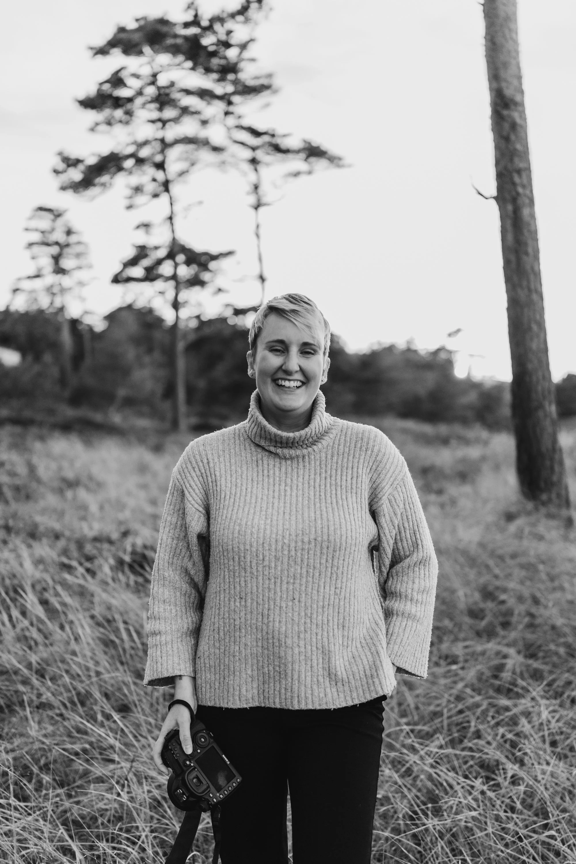 Bröllopsfotograf Denise Eriksson står med sin kamera i ett öppet gräslandskap med några träd i bakgrunden. Hon ler stort in i kameran. Bilden är svartvit.