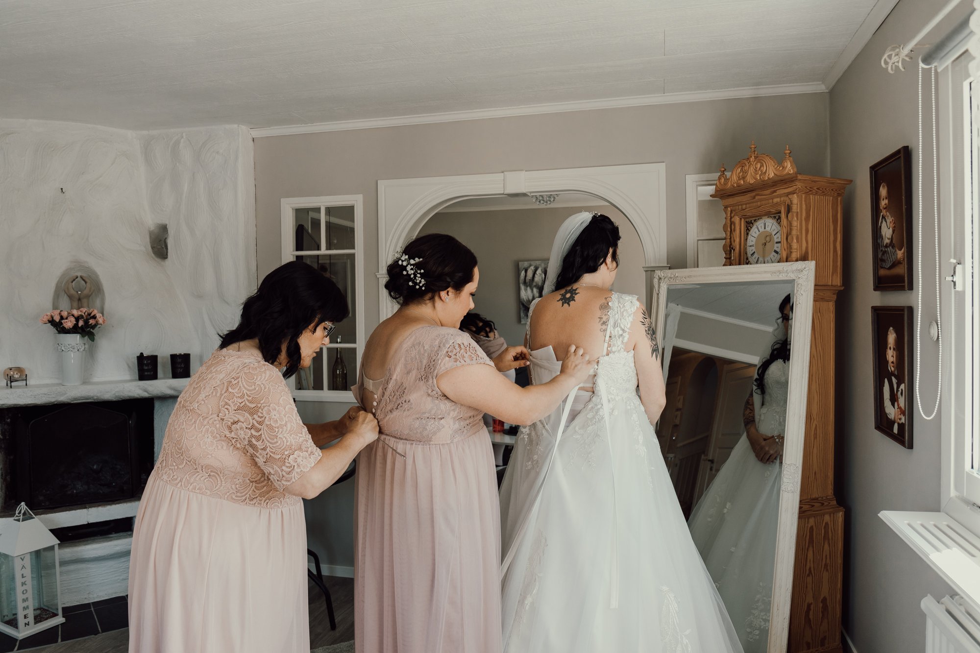 Ett led med tre kvinnor där bruden står längst fram med ryggen mot kameran, hennes syster står bakom och knäpper hennes klänning och modern står bakom systern och knäpper ihop en klänning.