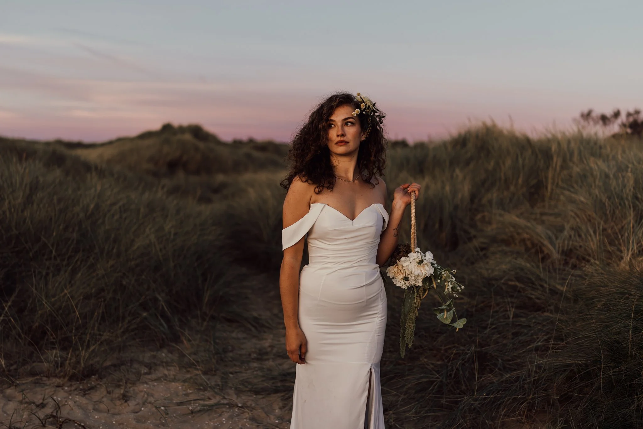 Brud står på en strand vid solnedgången och håller en korg med blommor.