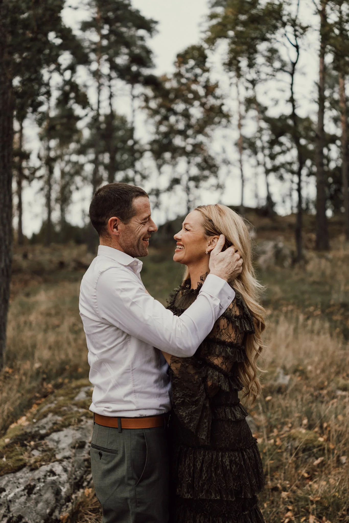 Par står tätt tillsammans i ett öppet landskap med högt gräs och träd i bakgrunden. Mannen håller varsamt om kvinnans ansikte medan de ler mot varandra.