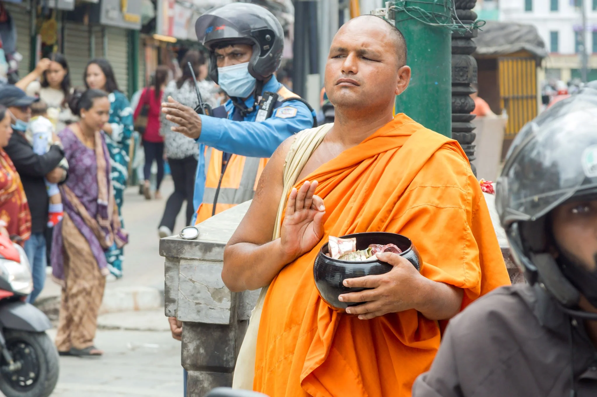A Buddhist monk maintains a serene meditative posture while collecting alms amid Kathmandu's chaotic traffic, one eye watchfully tracking the flow of vehicles around him.
