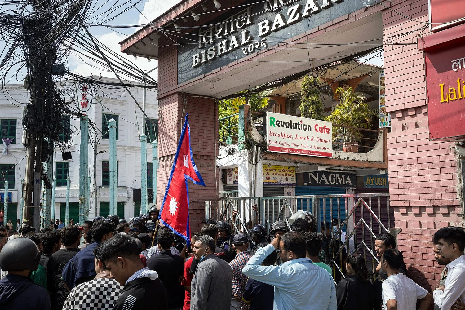 [Act II: Collision]. Crowds assemble in front of a police station at Bishal Bazaar, next to the Revolution Cafe.