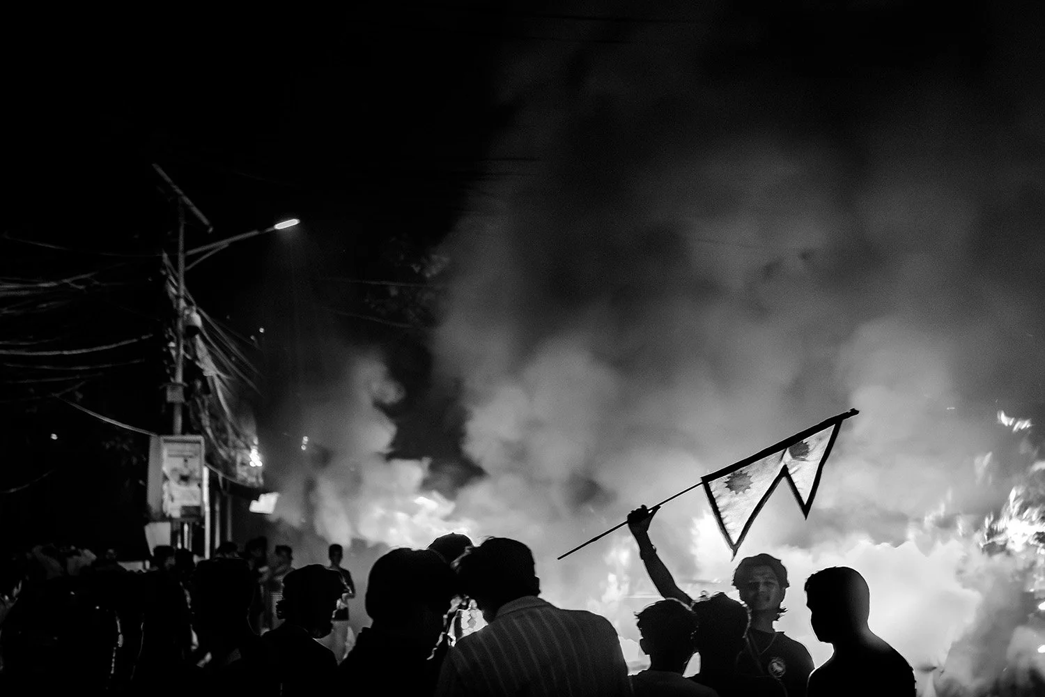 [Act II: Collision]. A protester raises the Nepali flag against a backdrop of fires