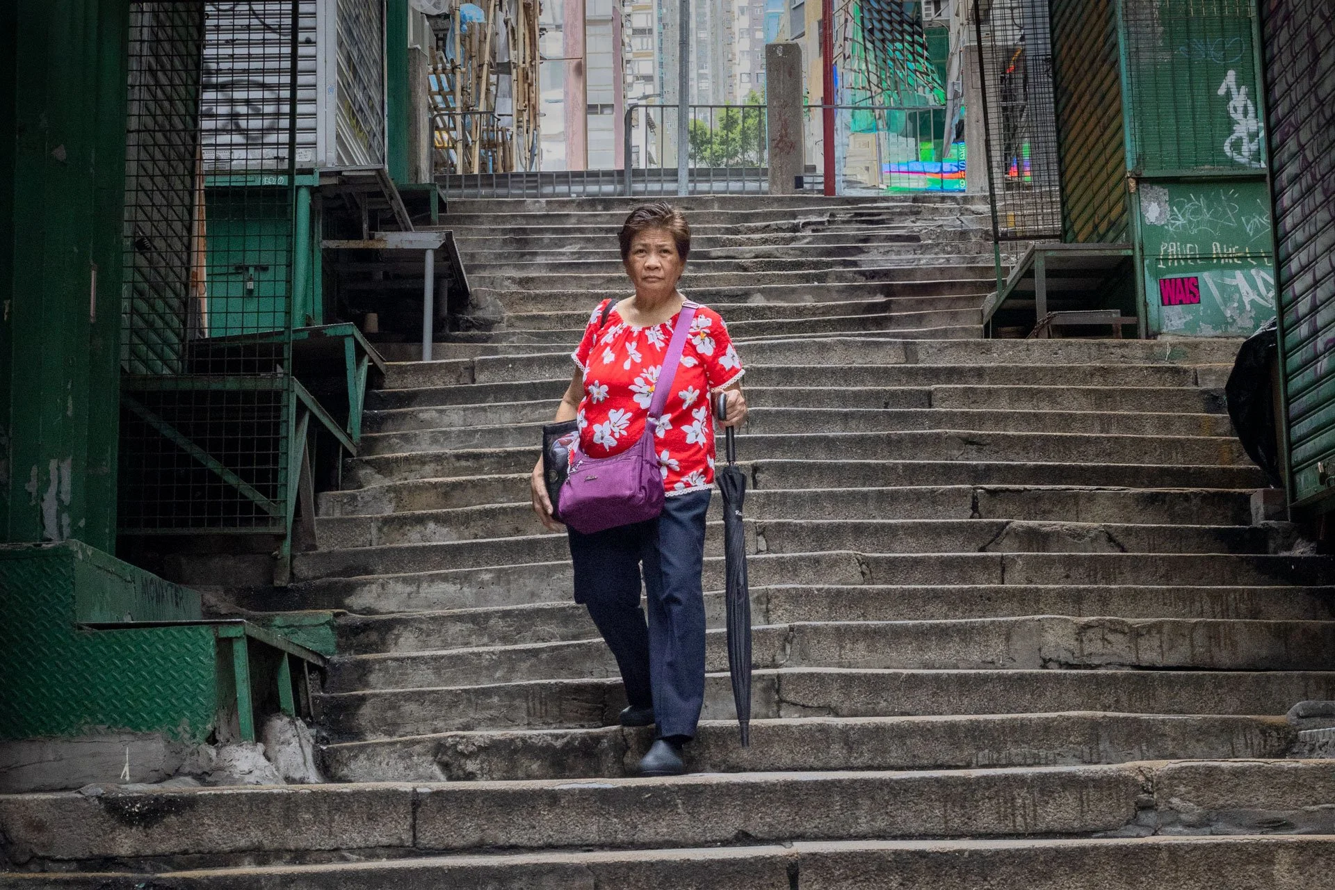 A woman in a vibrant red shirt descends stone steps through a narrow green-walled alley in Hong Kong, her bright clothing creating a striking contrast against the weathered urban passageway.