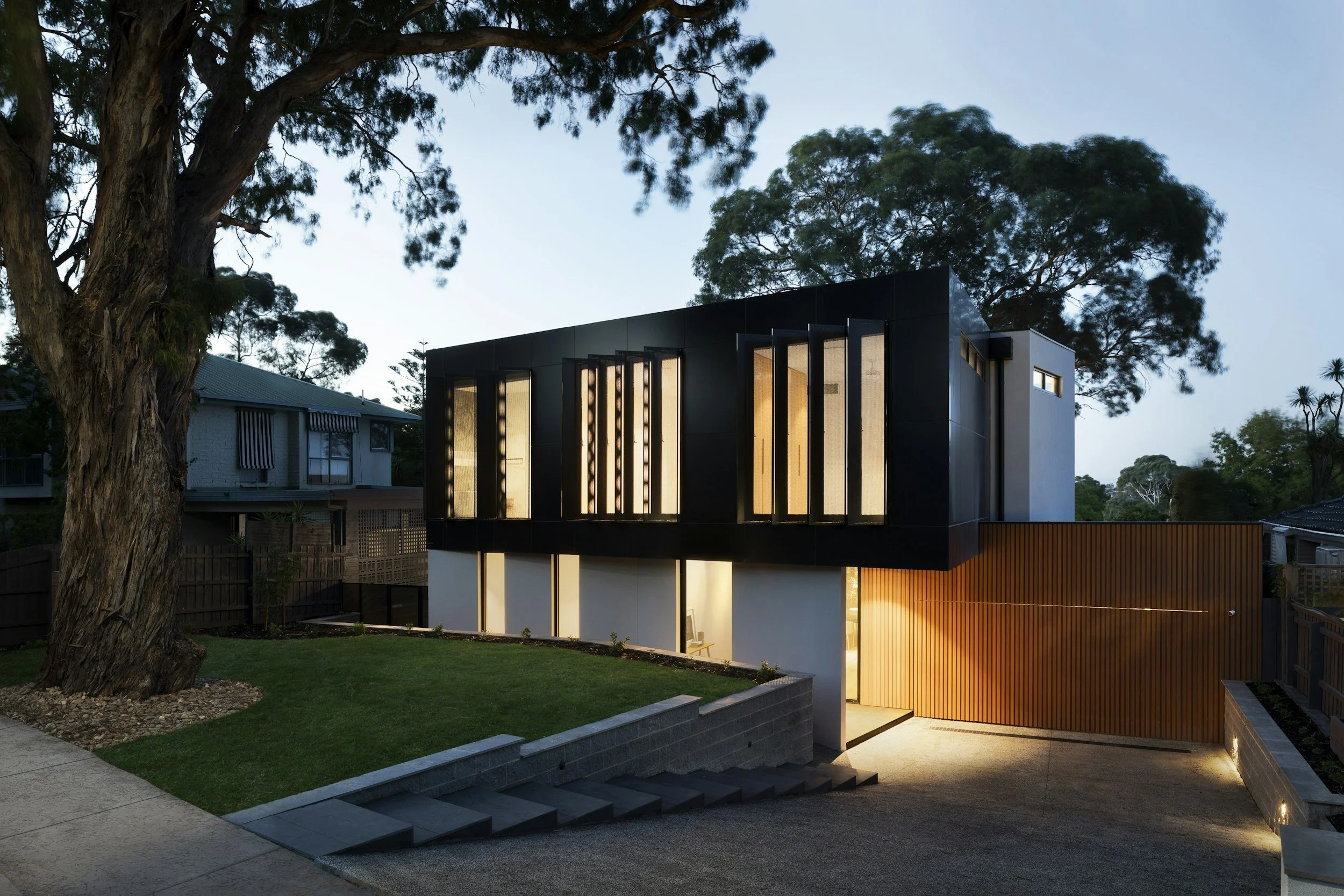 Modern two-story house with black and white exterior, illuminated windows, and wooden garage door, surrounded by trees and a landscaped yard during dusk.