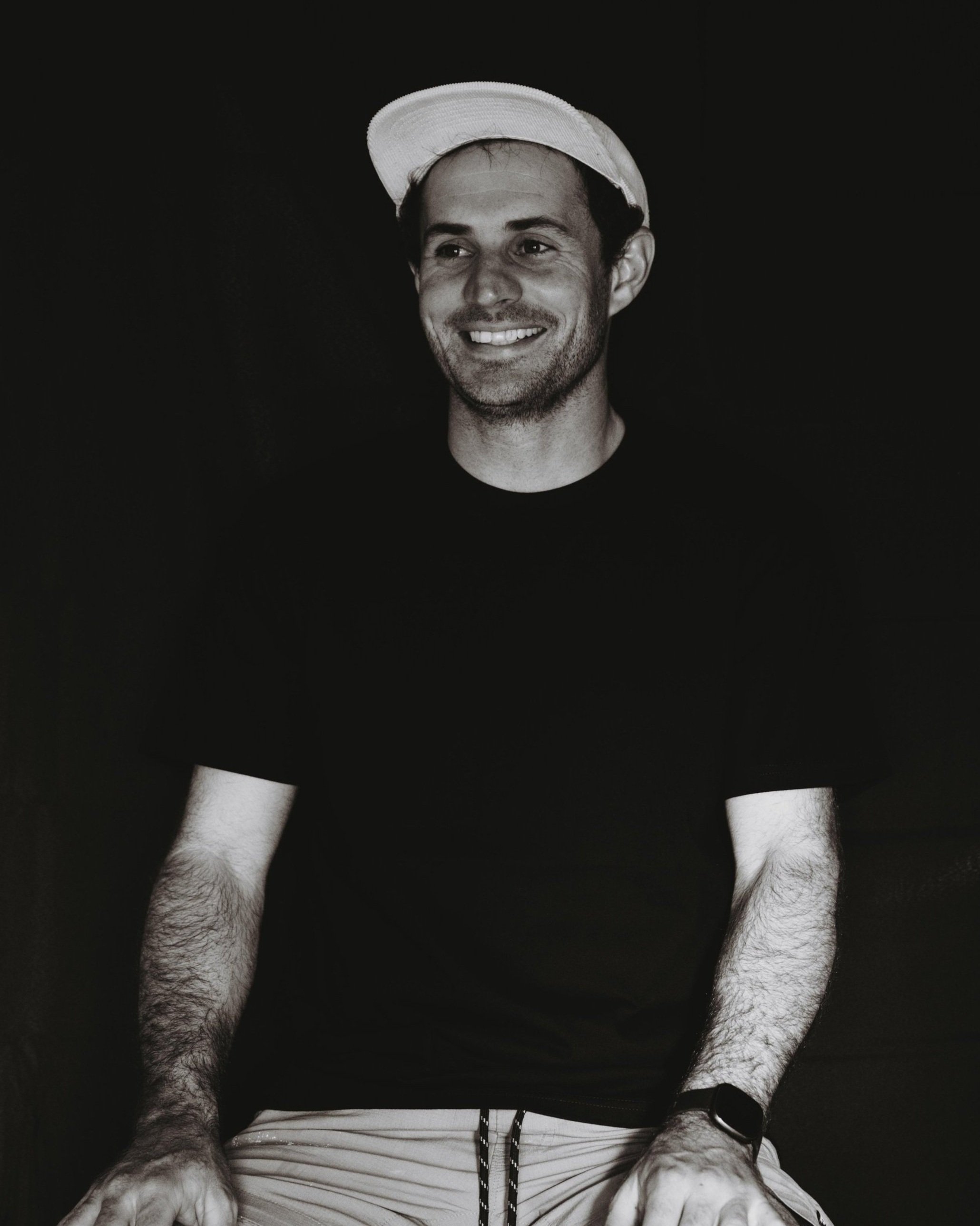 A young man with a smile, wearing a light-colored hat and a black t-shirt, sitting against a dark background.