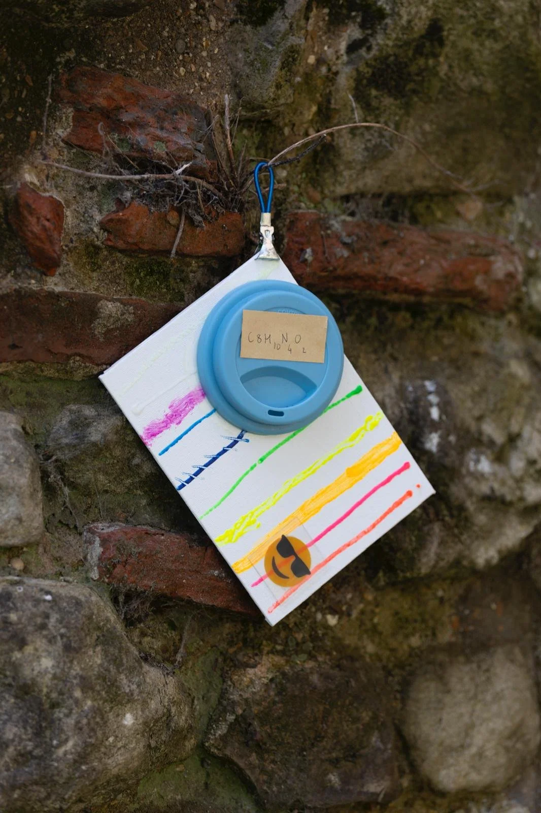 A piece of paper with rainbow-colored lines and a smiley face with sunglasses, hanging on a stone wall. A blue plastic container with a cardboard label that reads 'C8H10N4L' is attached to the paper, and a piece of string connects it to the wall.