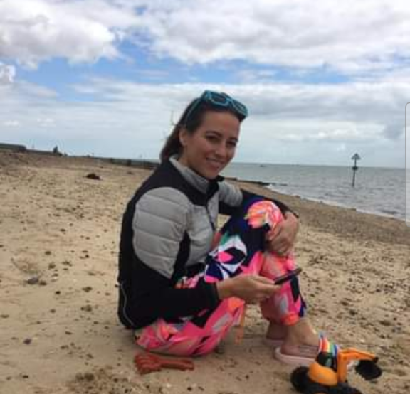 sitting on the beach, smiling. The woman is looking at a smartphone, wearing a black and gray jacket, colorful pants, and blue sunglasses. The girl is wearing pink, sitting with her legs crossed, and holding a toy; there are toys nearby, with the ocean and a cloudy sky in the background.