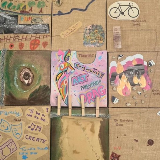 A collection of colorful, hand-drawn artwork and collages on a wooden surface, including a rainbow unicorn with the words "ART," "COMMUNITY," "FRIENDSHIP," and "DRAG," along with various other drawings and notes.