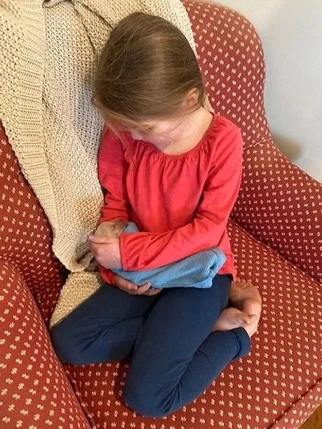 A young girl with long brown hair sitting cross-legged on a red patterned armchair, holding a blue blanket, wearing a red long sleeve shirt and dark pants, looking down.