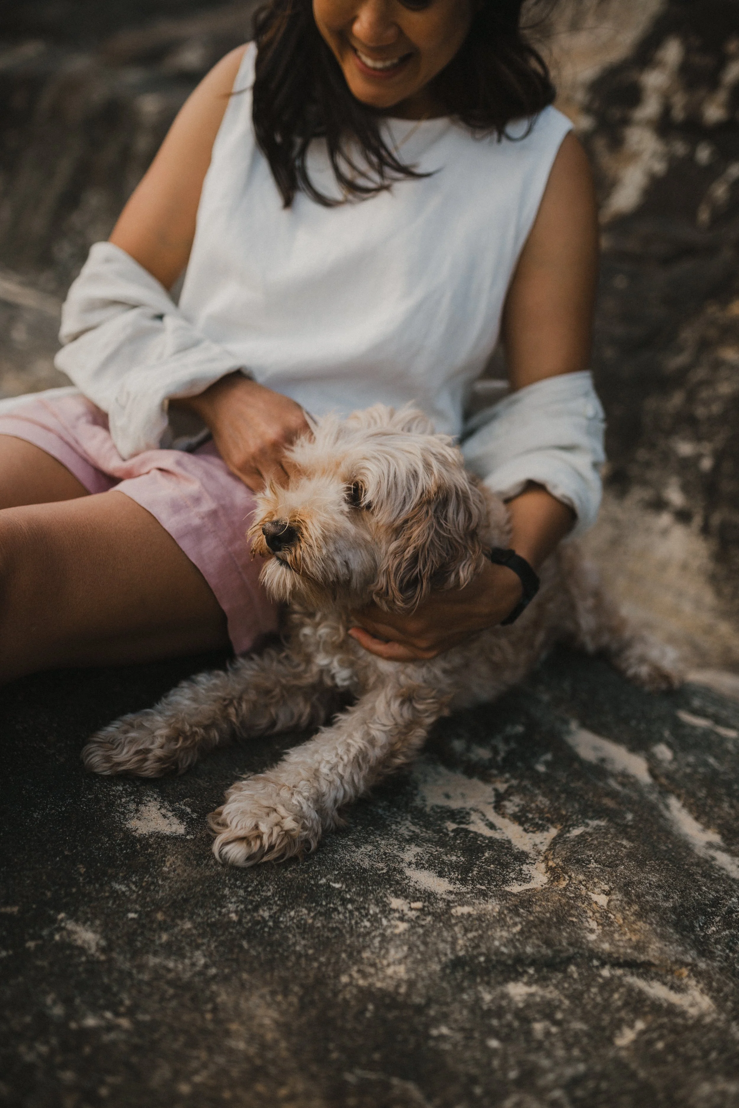 A woman sitting on a rock holding a small, curly-haired dog, smiling and looking down at the dog.