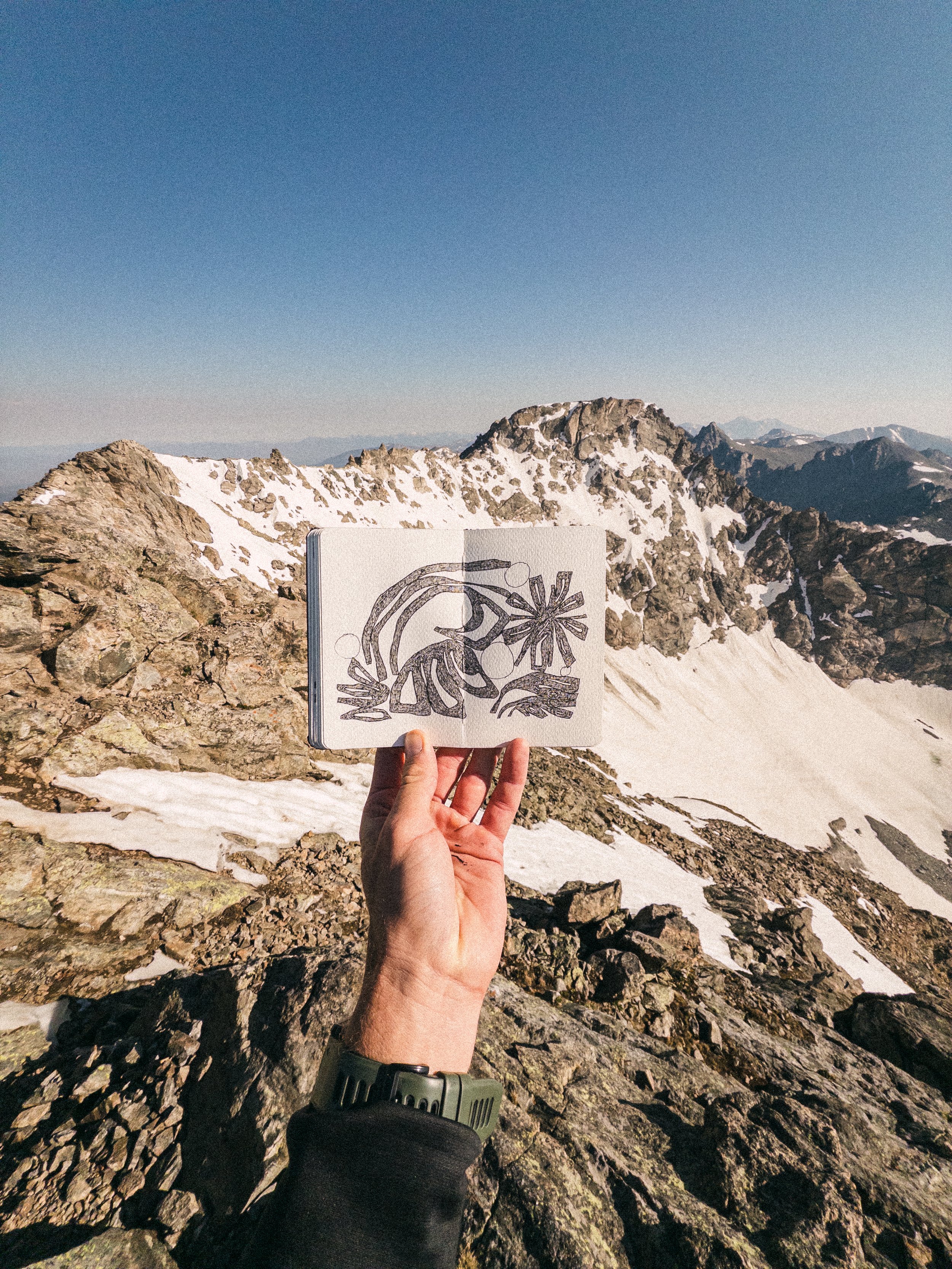 A person holding an open sketchbook with a drawing of a mountain landscape, standing on rocky terrain in a mountainous area with snow patches, under a clear blue sky.