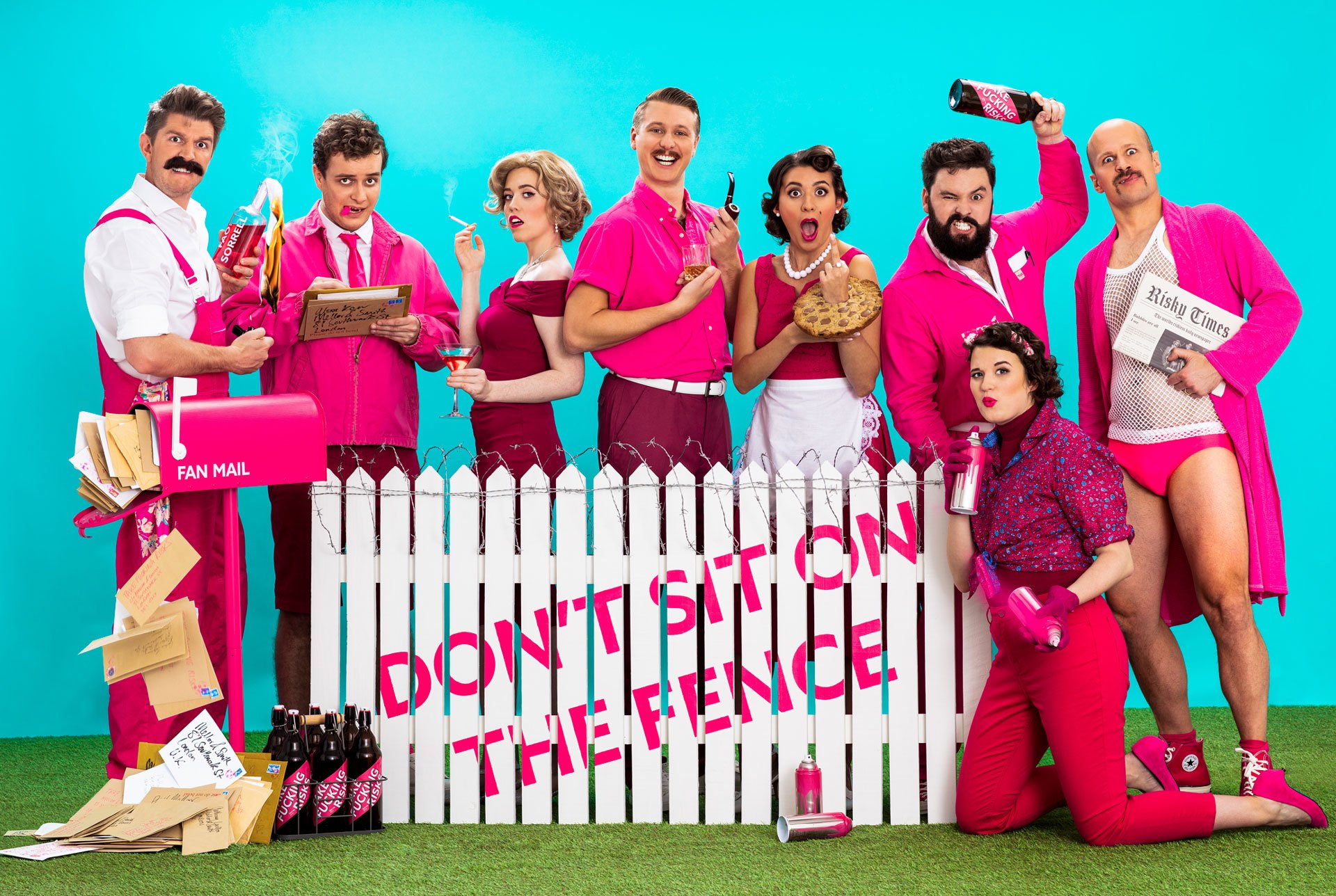 Group of people in colorful outfits posing humorously against a bright blue backdrop, standing and sitting around a white picket fence with the phrase "DON'T SIT ON THE FENCE." They are holding various props like fan mail, drinks, and a newspaper.