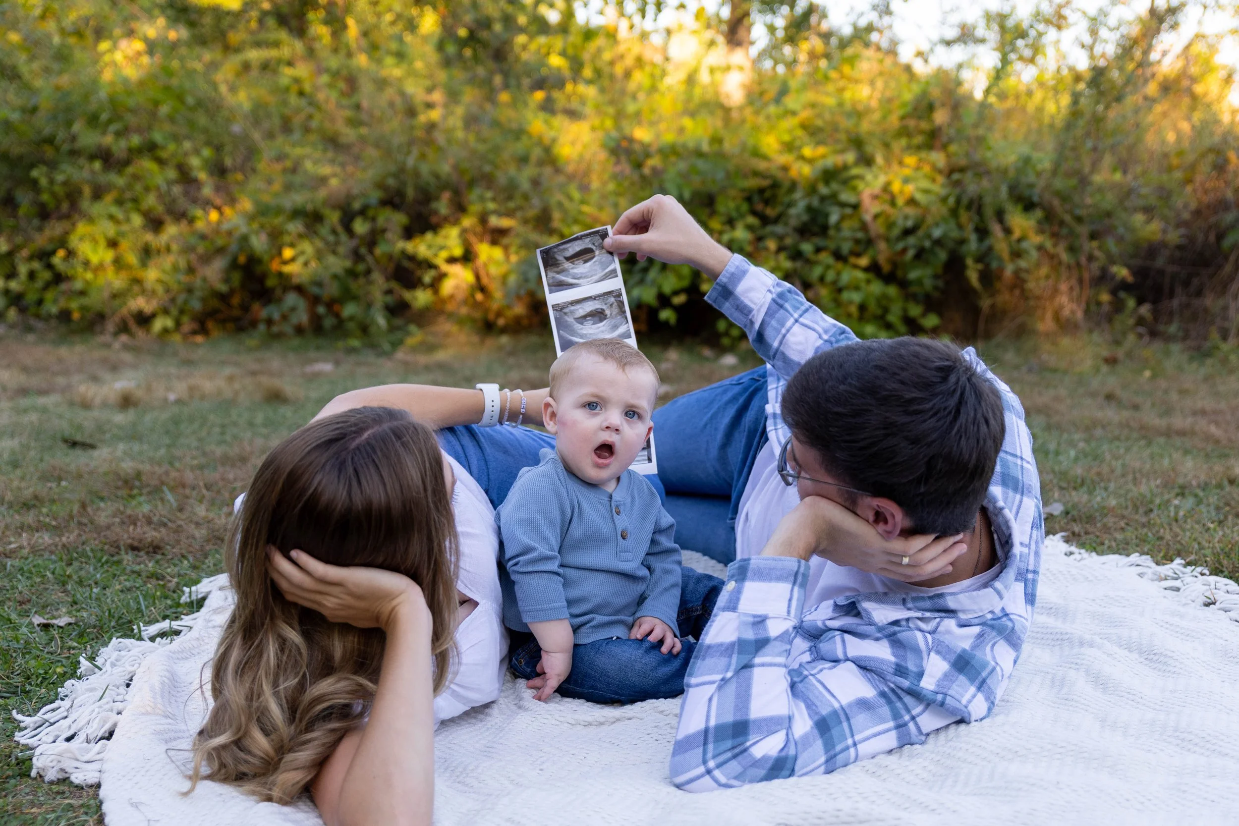 Family lying on a blanket outdoors, looking at ultrasound pictures, with a young child in the center and a man and woman on either side of him, in a grassy area with trees and bushes in the background.