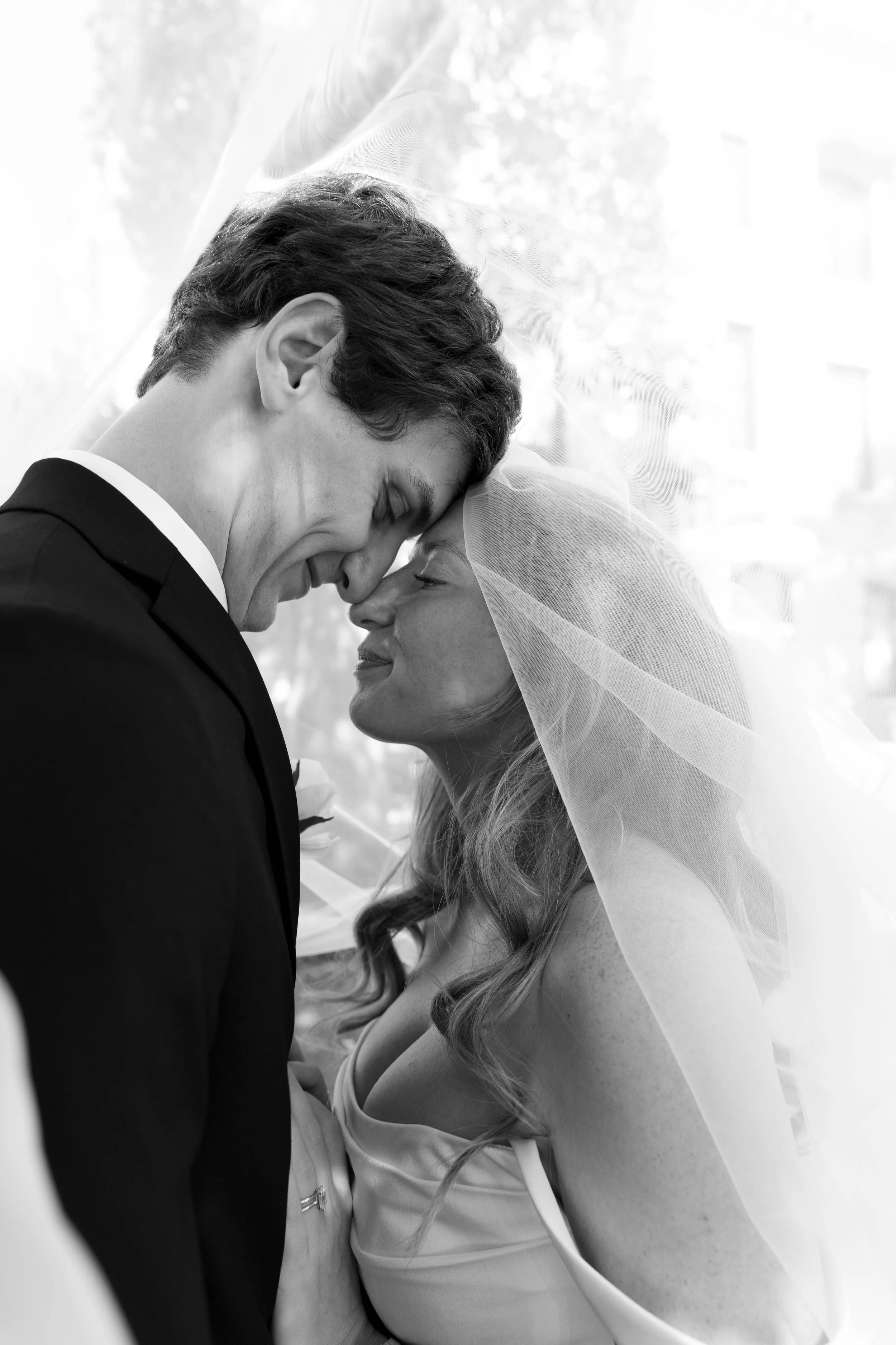 Black and white photo of a bride and groom with foreheads touching and eyes closed, smiling, during their wedding.