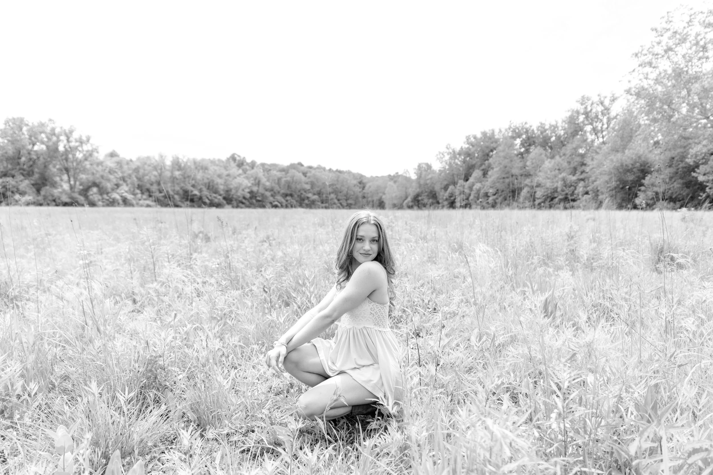 A woman with long hair, wearing a sleeveless dress, kneeling in a grassy field.