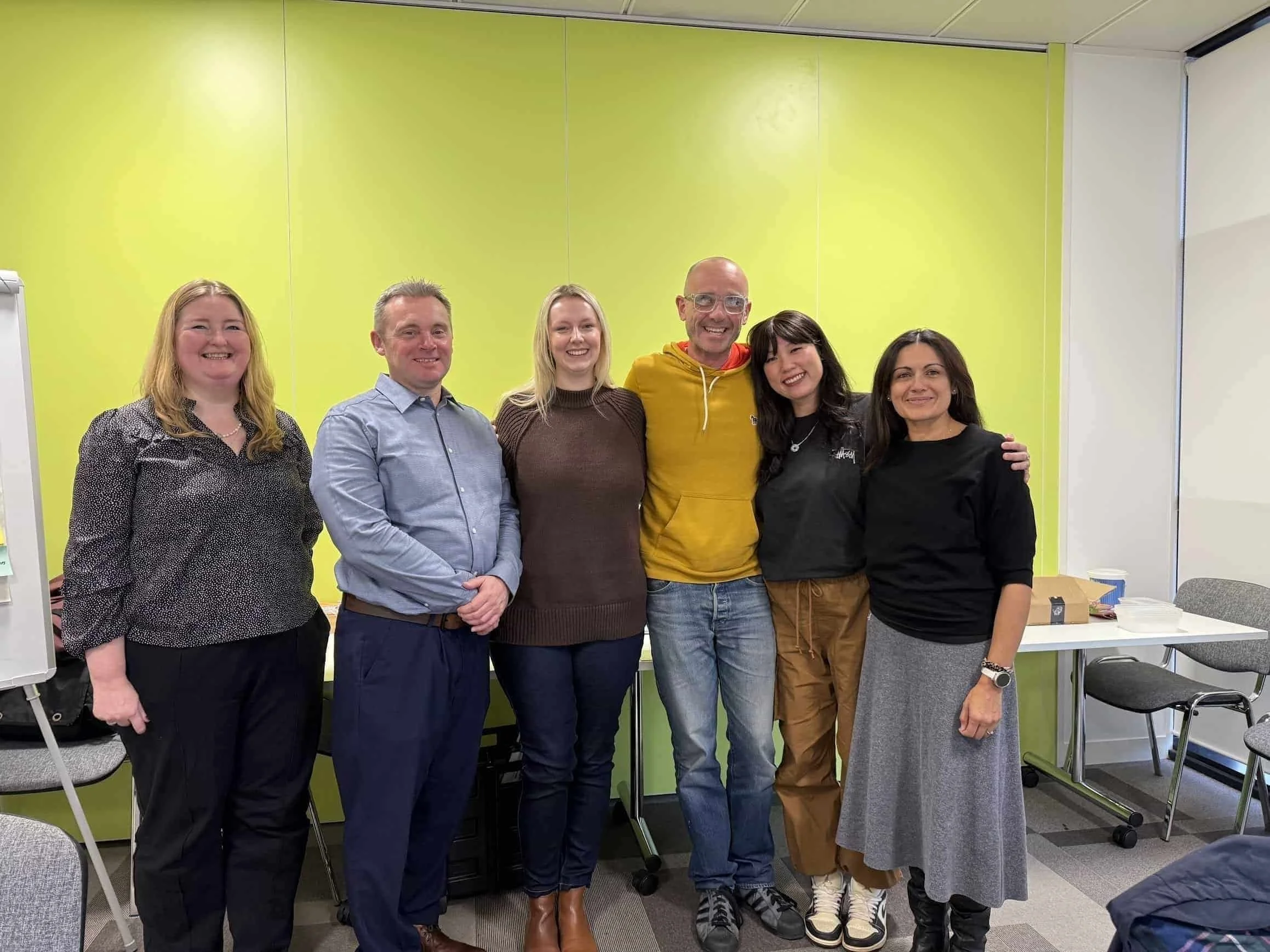 Six men and women stood in front of a green wall in an office environment. They are all dressed casually, looking at the camera smiling