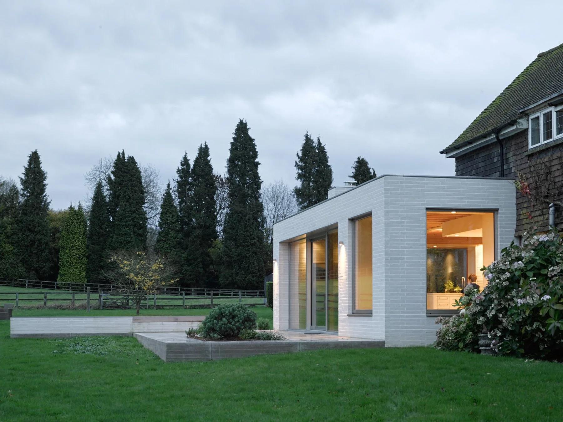 Modern white brick house with large windows and sliding glass door, illuminated from inside, situated in a lush green yard with trees and bushes, under a cloudy sky.