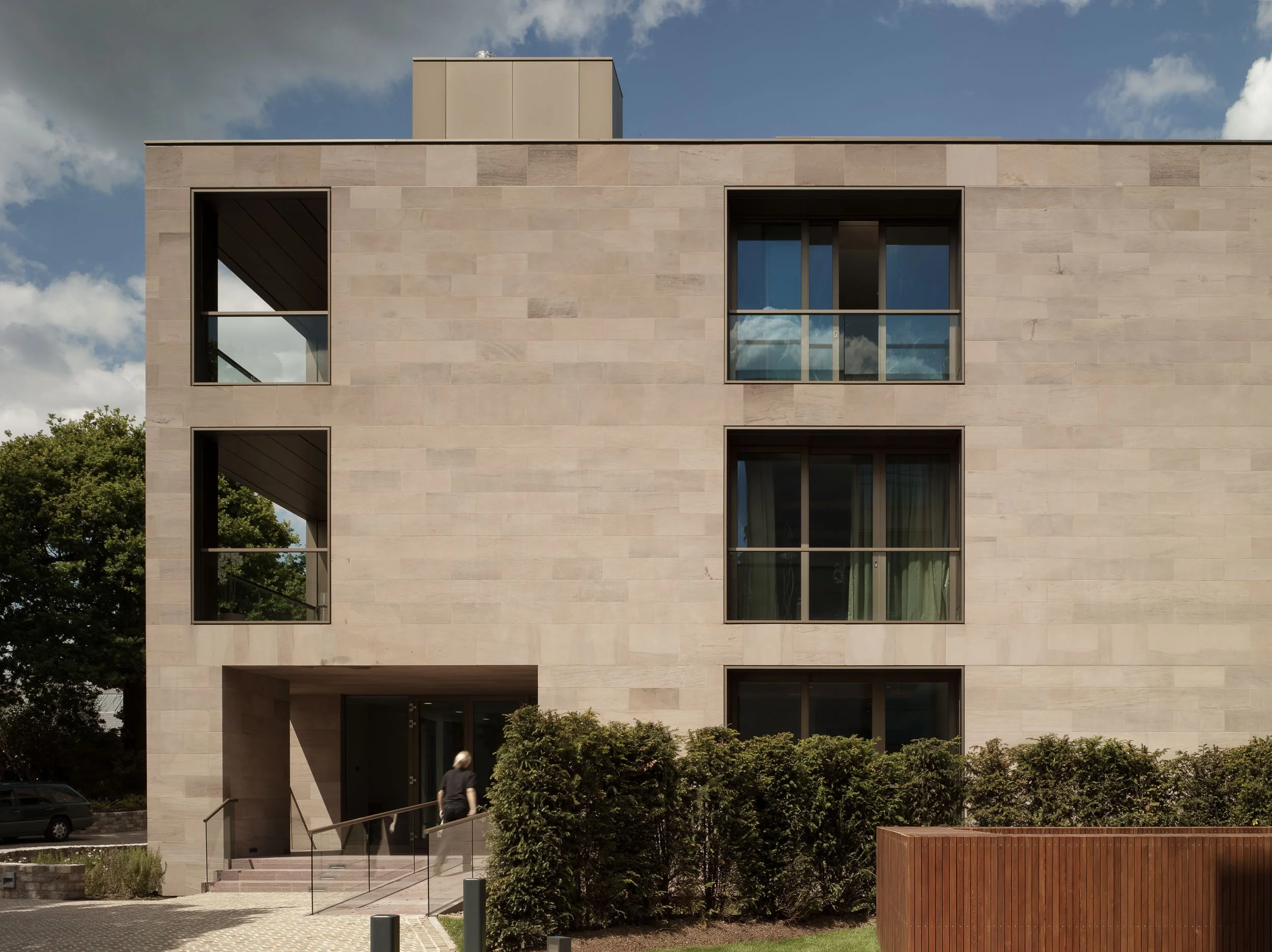 Modern beige building with large rectangular windows, there is a person walking at the entrance, green bushes and trees in front, and a partly cloudy sky in the background.