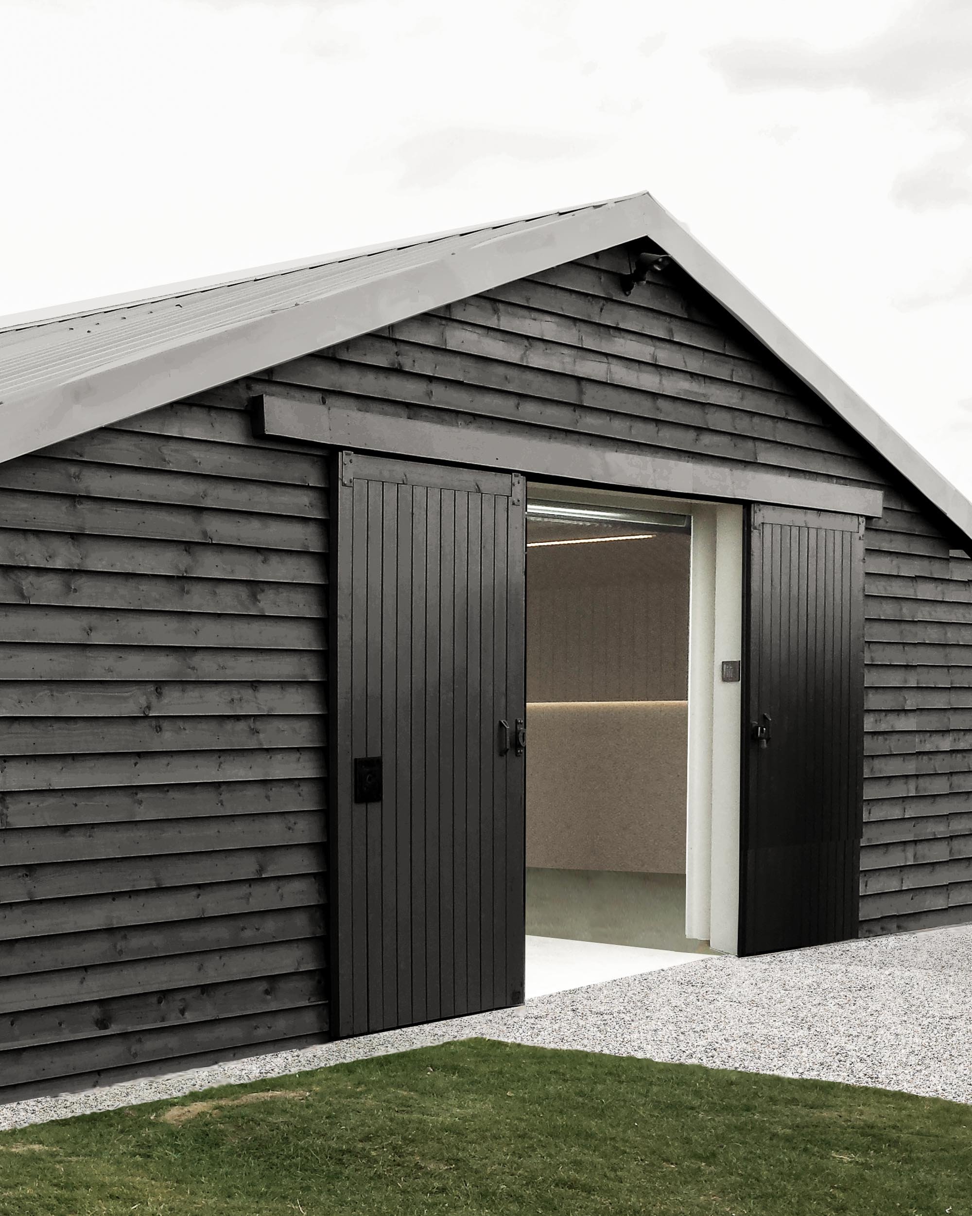A wooden building with dark gray vertical shutters on the windows, an open door revealing a hallway, and a sloped metal roof with a spotlight attached underneath. The building is surrounded by a patch of green grass and gravel pathway.