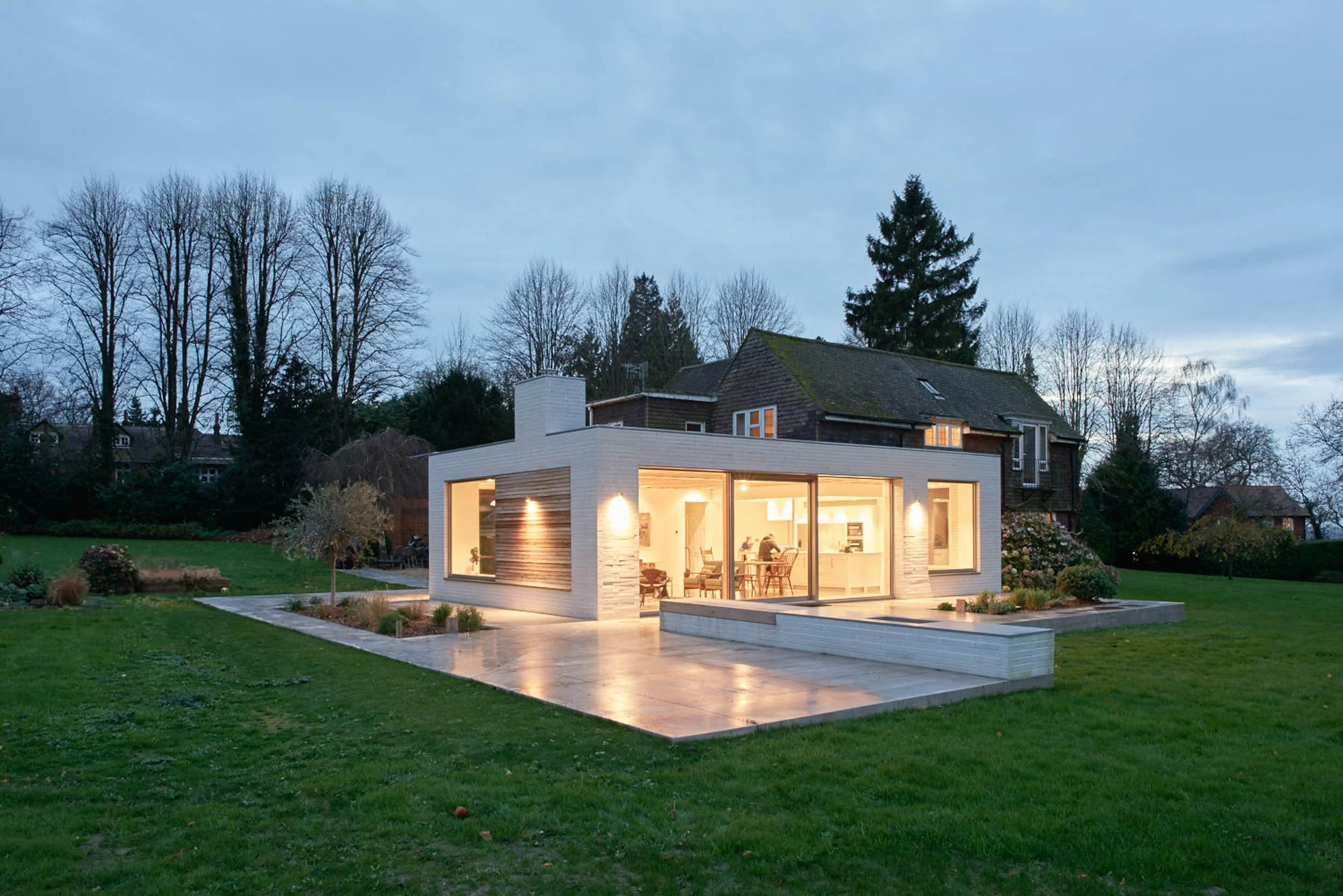 A modern single-story house with large glass windows emitting warm light, situated on a green lawn with landscaping, in a neighborhood with older houses and tall trees, during dusk or early evening.