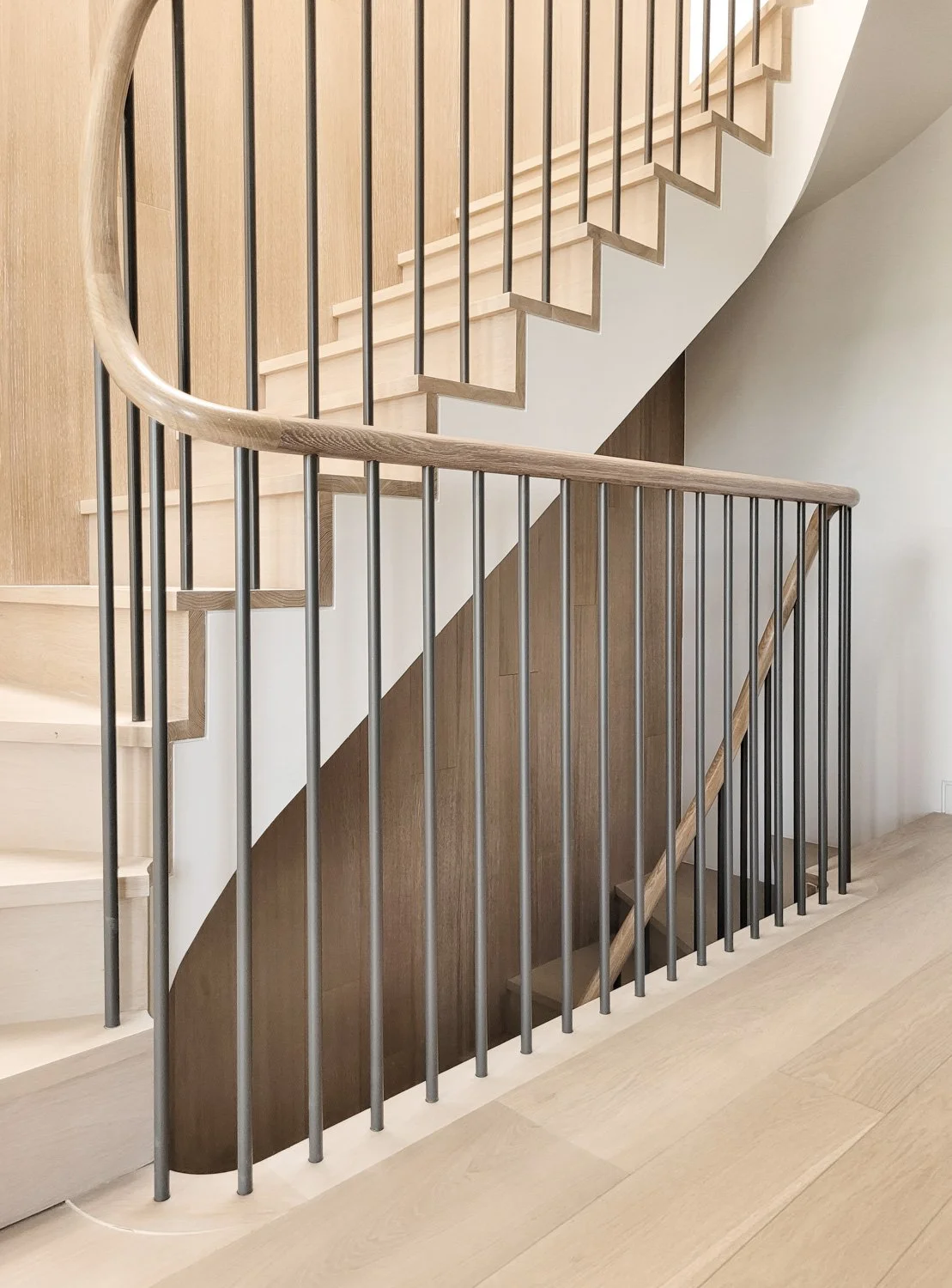 Interior view of a modern staircase with light wooden steps, black metal balusters, and a light wooden handrail, featuring a curved design.