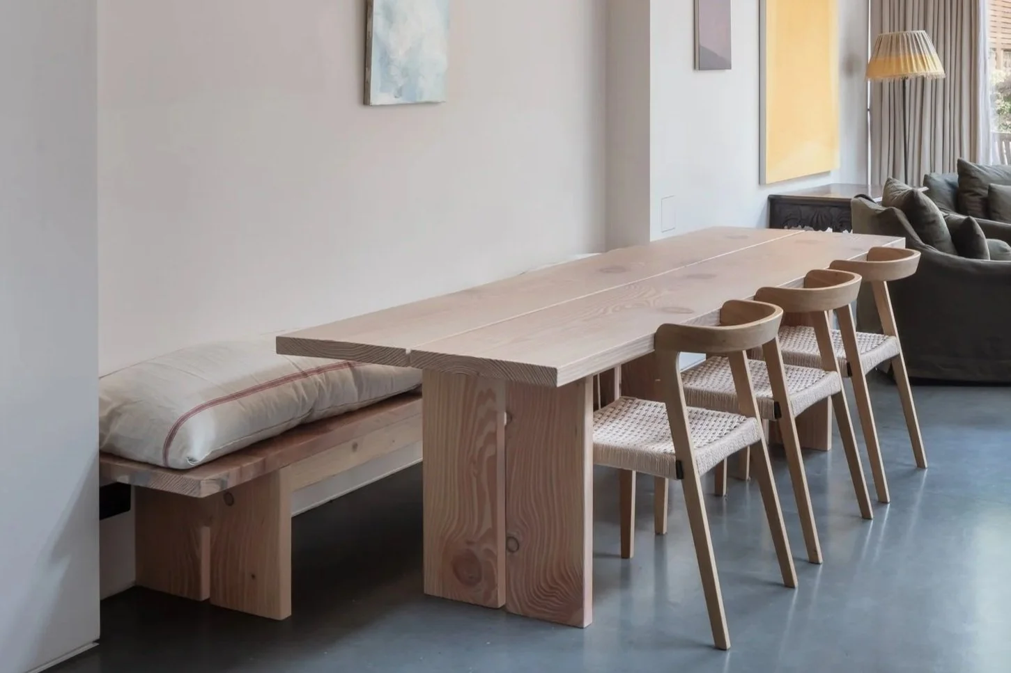 Minimalist dining area with a wooden table, four chairs with woven seats, a built-in bench with a pillow, and abstract wall art.