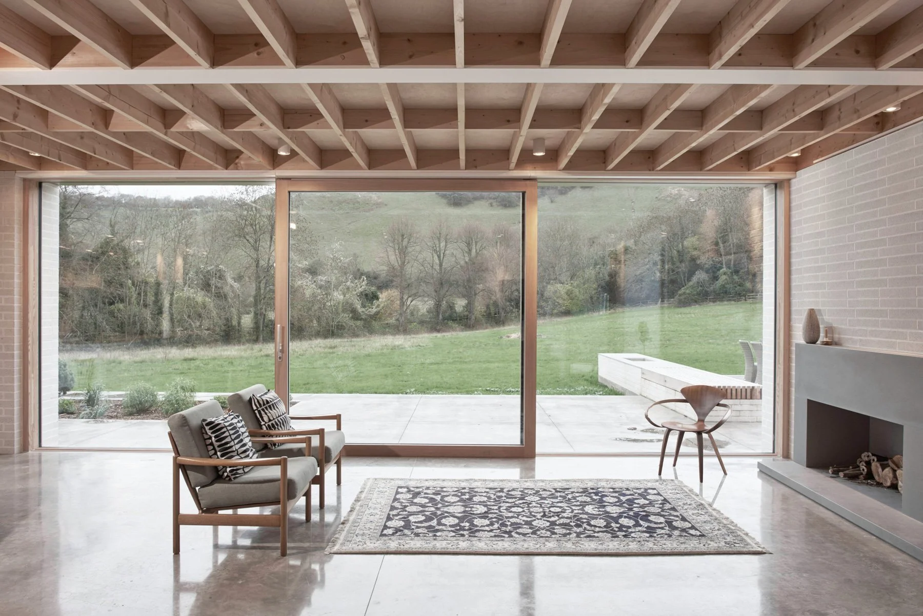 Modern living room with large floor-to-ceiling glass windows showing a view of green landscape and trees outside. The room features a fireplace, a patterned area rug, two chairs with patterned cushions, and a wooden chair near the window.