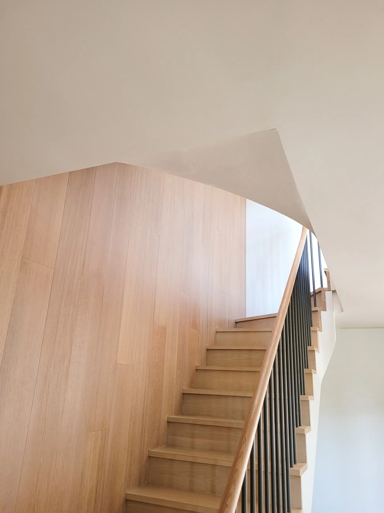 Wooden staircase with black metal railing next to a textured wall, leading to an upper floor with a small landing area.